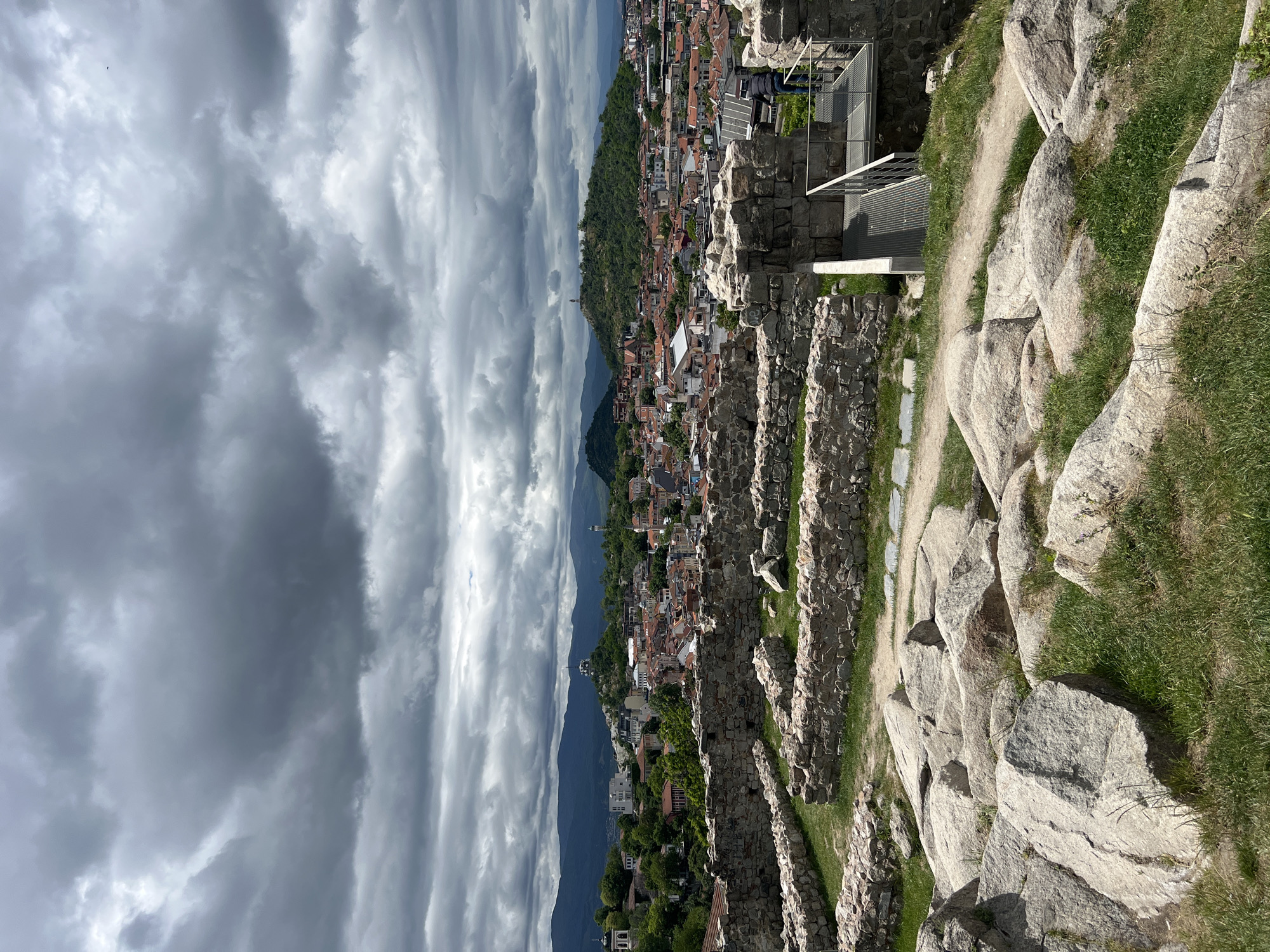 The image presents a view from Nebet Tepe, one of the hills in Plovdiv, Bulgaria, overlooking the city. The sky is overcast with dark, striated clouds. In the foreground are stone ruins, with patches of grass interspersed. A metal walkway with railings is visible. Beyond the ruins, the city of Plovdiv stretches out, with buildings and trees covering the landscape. Mountains are visible in the distance.