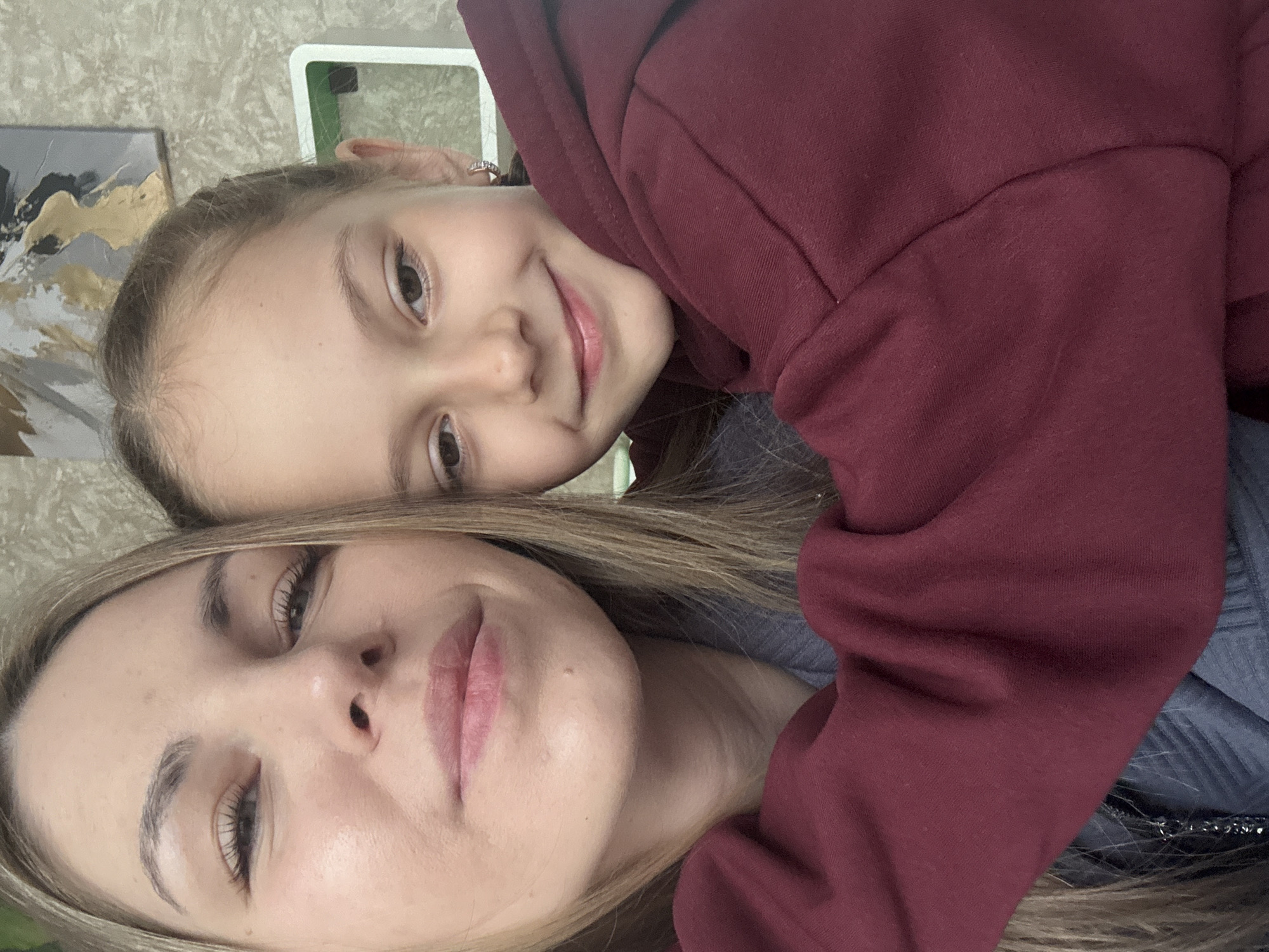 A selfie-style close-up of a woman and a girl. The girl has brown hair pulled back, brown eyes, and is smiling. She is wearing a maroon hoodie. The woman has blonde hair and is wearing a blue top. The background includes a beige patterned wall and a white shelf. The photo was likely taken indoors. The location in the city of Раздолье, Russia is not visually evident.