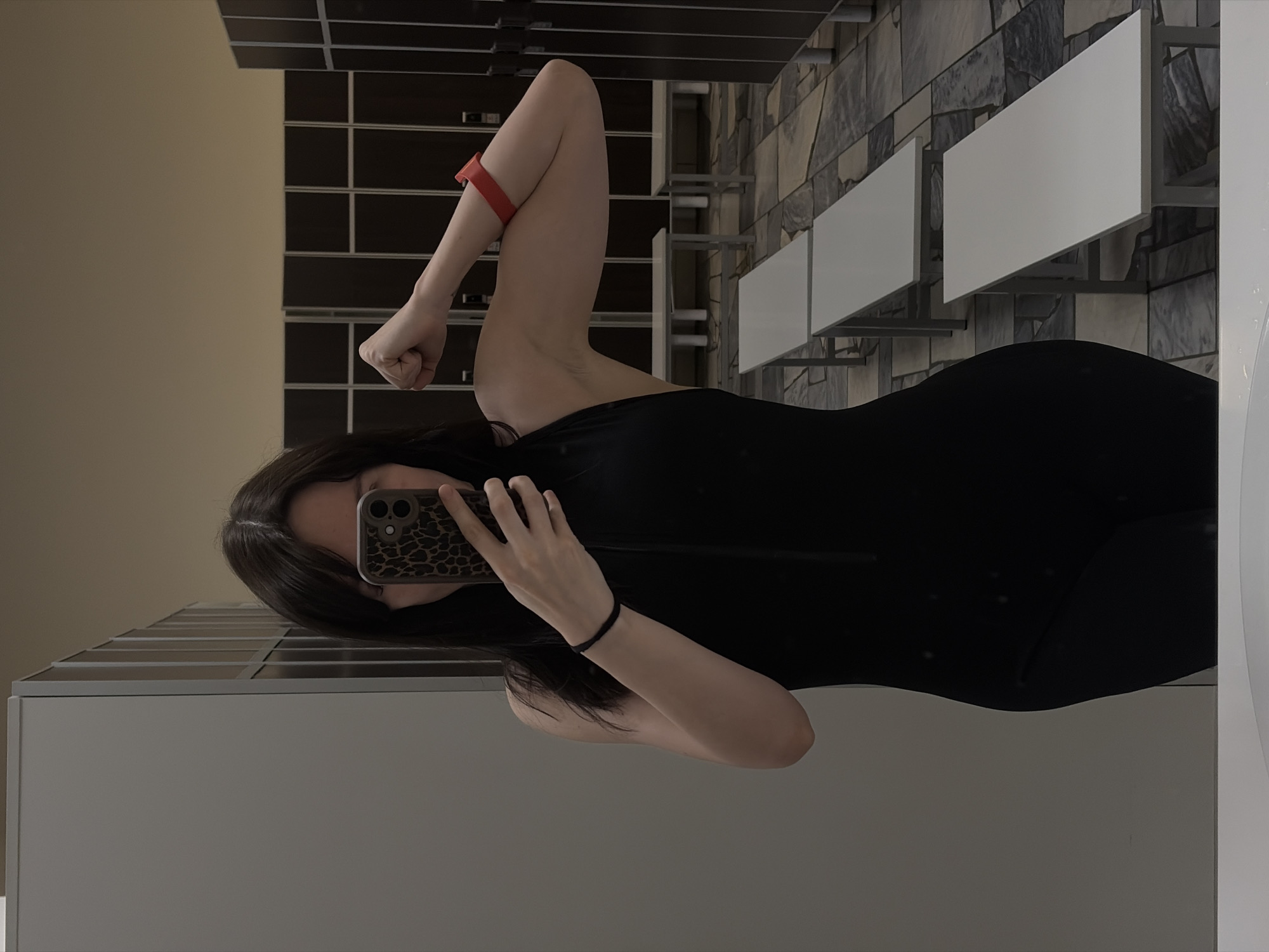 A young woman is captured in a mirror selfie within an indoor facility, likely a gym or changing room, potentially located in Chaikovsky, Russia. She is wearing a black form-fitting garment and has long, dark hair. Her face is partially obscured by a smartphone with a distinctive leopard print case, which she holds in her left hand.
The woman is striking a pose, flexing her right arm to showcase her bicep. A red elastic band is visible on her upper right arm, and a black elastic band adorns her left wrist. The background features dark brown lockers or storage units with silver handles on the left. To the right, a wall is covered with grey and brown patterned tiles, upon which several white, rectangular tables or shelves are mounted with metal supports. A light-colored wall or reflective surface occupies the immediate foreground on the left, and the bottom part of the image shows a reflection, indicating the selfie was taken in a mirror. The lighting is ambient, characteristic of an indoor environment, making the time of day indeterminate. No discernible text is visible within the image. Her pose conveys a sense of confidence or pride in her physical state.
