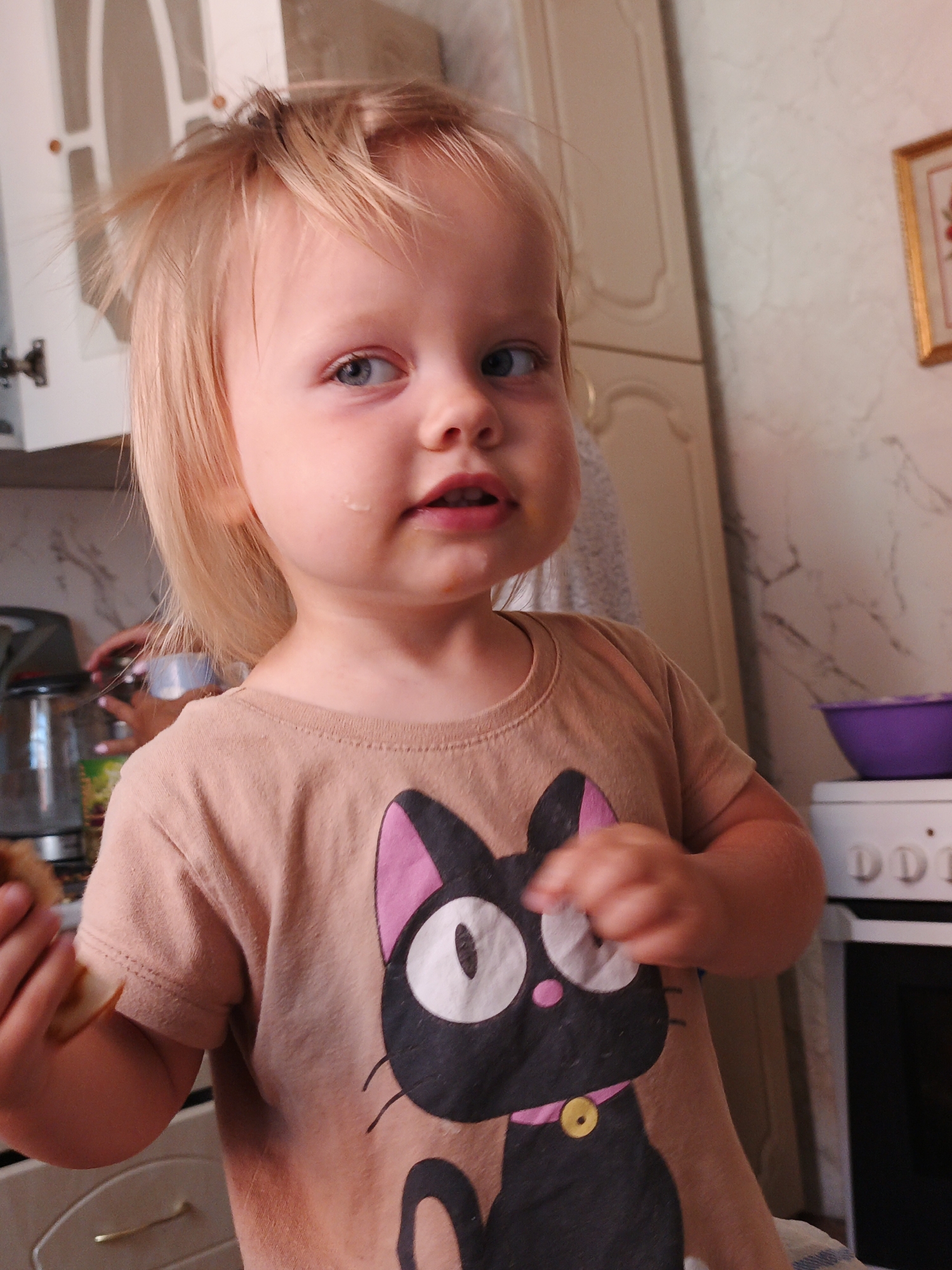 A close-up, eye-level shot shows a young Caucasian child with blonde hair and blue eyes holding a piece of bread near their mouth. The child is wearing a tan t-shirt with a large black and pink cat graphic. The background is blurred but appears to be a kitchen with white cabinetry and appliances. A framed picture with a red floral motif is visible on the wall to the right. The child's left hand is visible in the foreground, holding the bread. The overall lighting is soft, and the focus is on the child's face and torso. The image was captured in Medvedyovka, Russia.