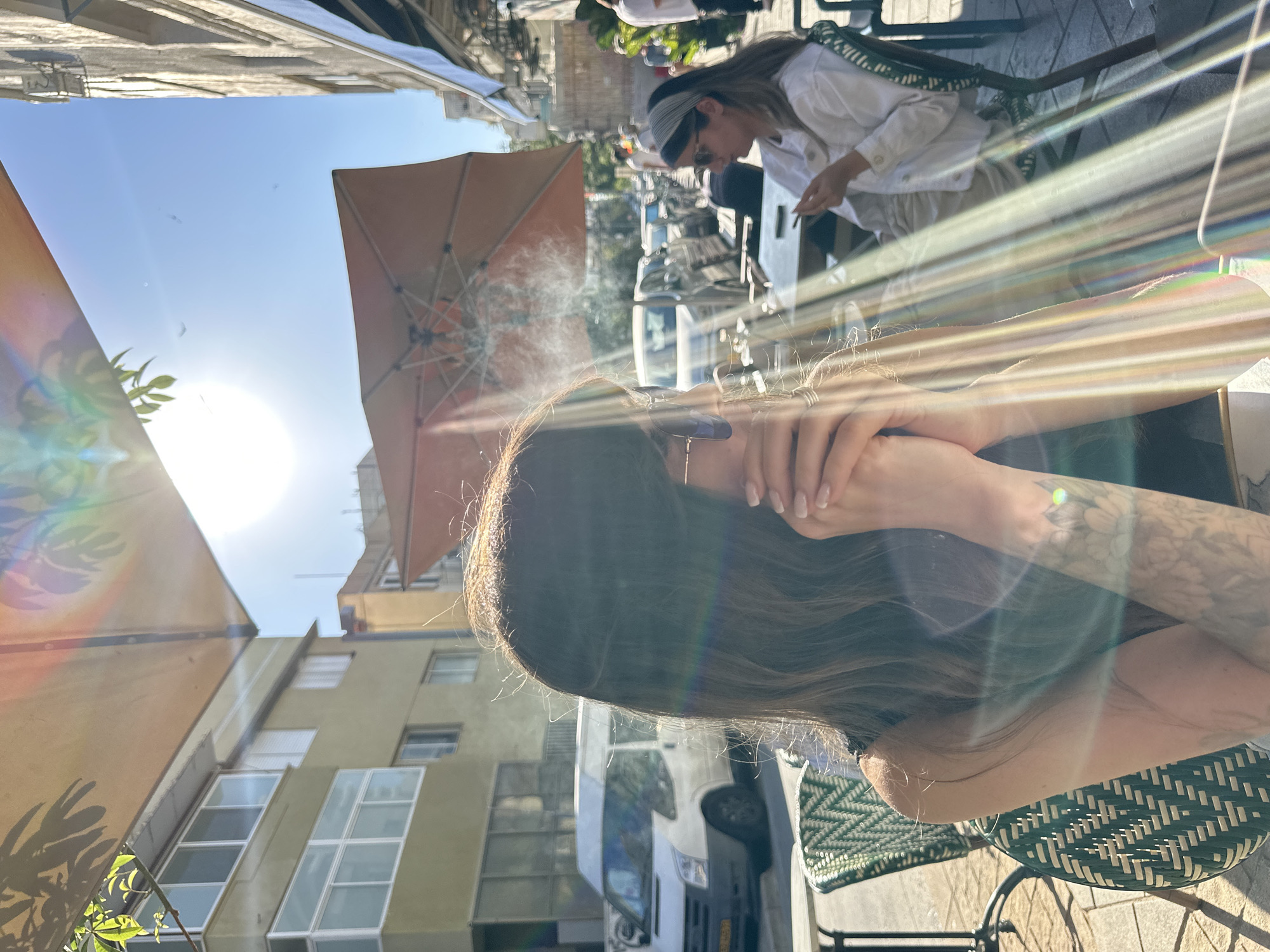 The image appears to be taken at an outdoor café in Tel-Aviv, Israel. The camera is positioned low, looking upwards. A person with long brown hair and a tattoo on their arm sits at a woven chair. Their hands are held up near their face. Above them, another person with a headband and sunglasses sits at a table, possibly smoking. A large orange umbrella provides shade. In the background, there's a street with parked cars and buildings characteristic of Tel-Aviv, including light-colored buildings with numerous windows. The sky is bright blue with strong sunlight creating lens flares. There are wisps of smoke in the air near the umbrella.