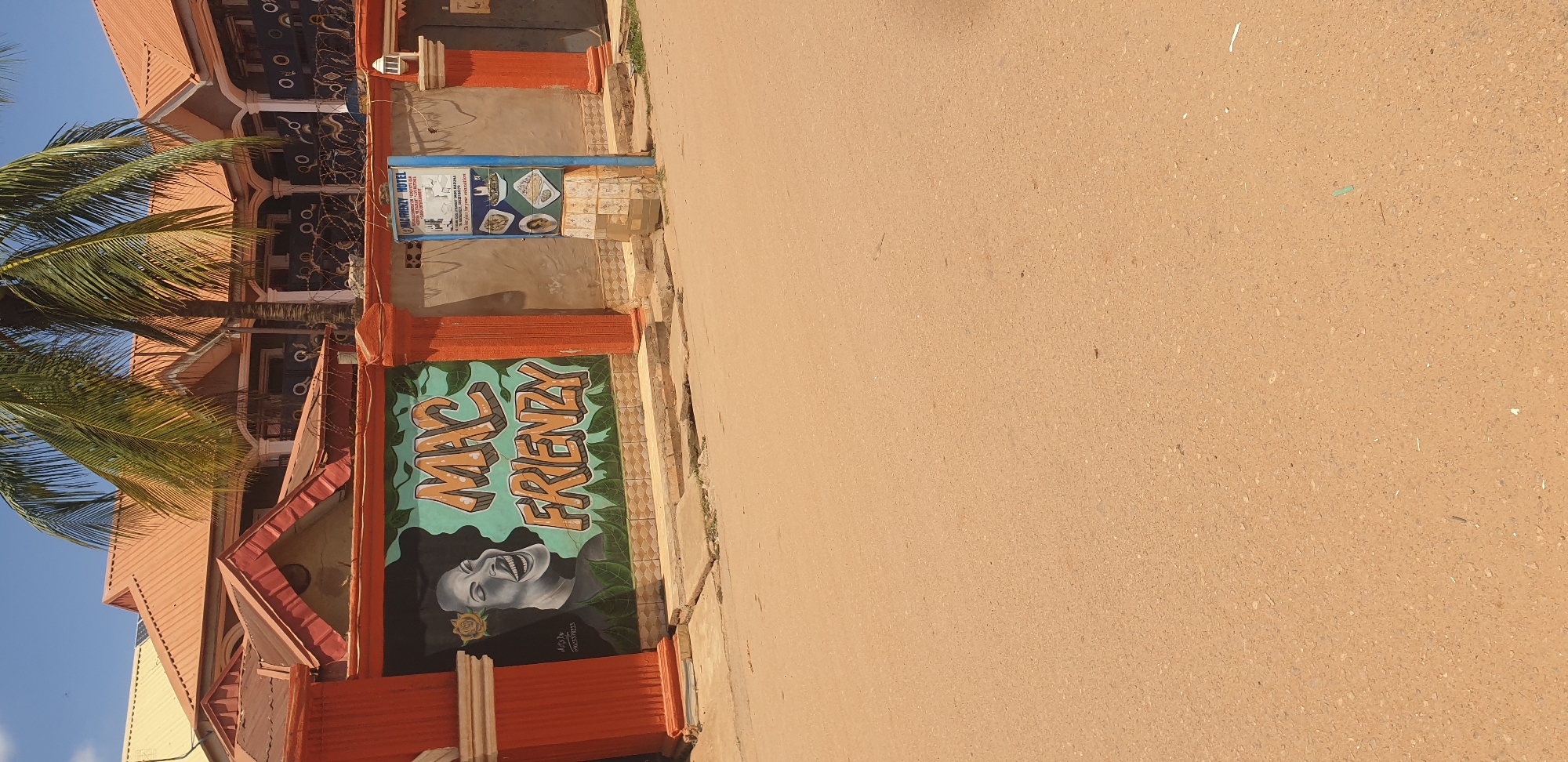 This image depicts an exterior view of the "MAC FRIENZY HOTEL" in Sabon Tasha, Nigeria, under a clear daytime sky. The foreground consists of an unpaved, light brown-tan road with a fine gravel texture, extending from the bottom of the frame upwards. A low concrete curb separates the road from a perimeter wall.
The wall, painted in an orange-red hue with decorative columns, features a large mural. On the left side of the mural, a grayscale depiction of a smiling woman with a yellow rose in her dark hair is visible. To the right of the woman, the words "MAC FRIENZY" are rendered in a three-dimensional, orange-brown graffiti-style font with green outlines, set against a light blue-green background decorated with dark green leaf patterns. Barbed wire runs along the top of the wall.
To the right of the mural, a freestanding blue sign post displays "MAC FRIENZY HOTEL" at the top. Below, a list of services such as "GOOD ACCOMMODATION," "SECURITY," "RESTAURANT," "BAR," "LIVE MATCHES," and "CYBER ENTERTAINMENT" is presented in white text. The sign also features small photographic images of food dishes and text stating, "The best place for your relaxation."
Behind the wall and sign, the multi-story hotel building is visible. It features a reddish-brown corrugated metal roof with multiple gables and a light-colored facade with white support pillars and dark-colored railings or decorative elements on the balconies. A large palm tree stands prominently in front of the building, its fronds extending into the upper right portion of the frame. No individuals or active subjects are present in the scene.