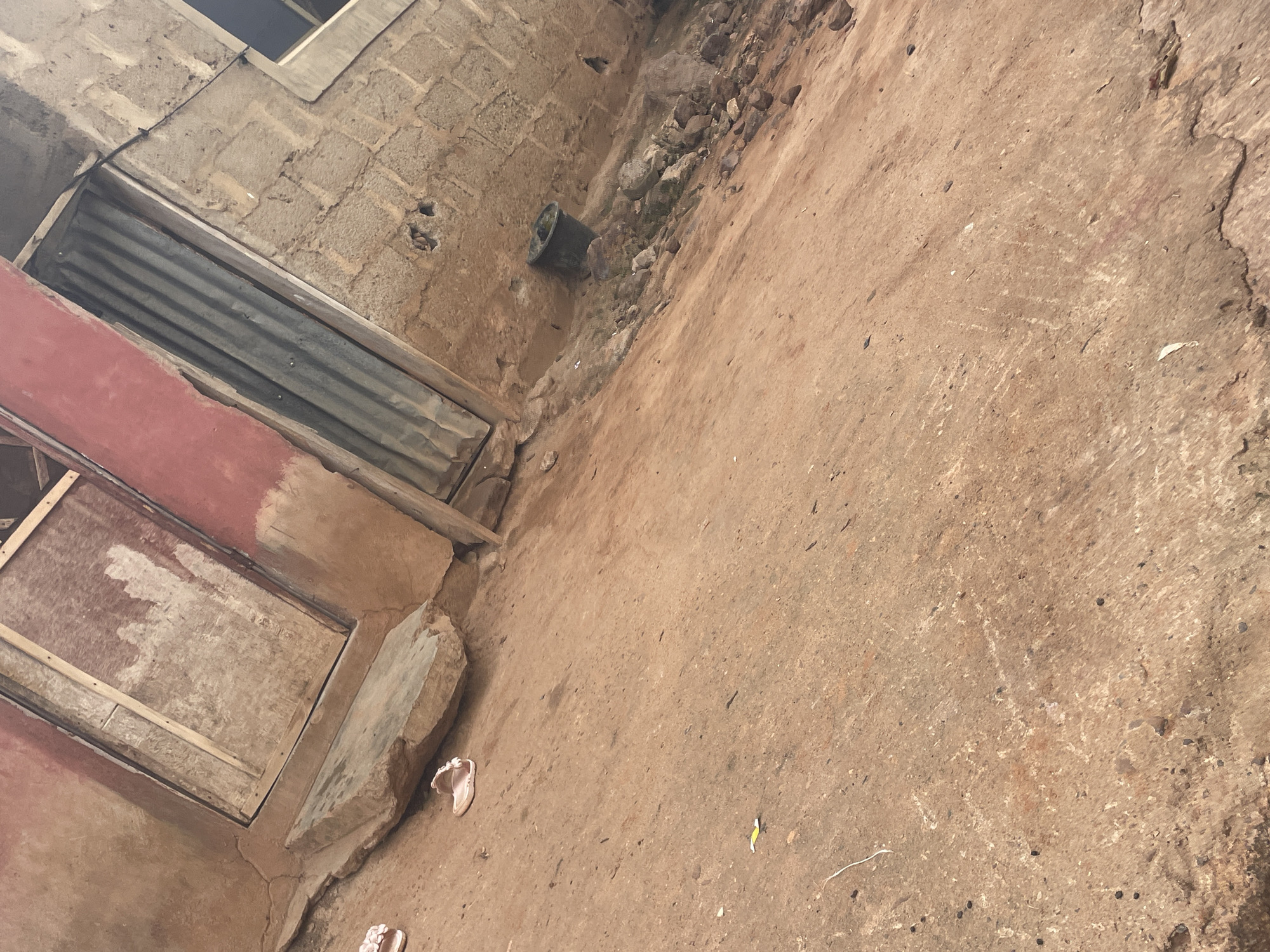 The image shows an outdoor scene in Lokoja, Nigeria, featuring the exterior of a building with rough, textured walls made of cement blocks. In the foreground, a light brown, dirt-covered ground is visible, with scattered small stones and debris. Two pairs of pink sandals are on the ground, one near a large, flat rock which serves as a step or platform. The building has two doorways; one appears to be a wooden door with a weathered surface, and the other is a corrugated metal door. Adjacent to the metal door, a dark cylindrical object, possibly a bucket, rests on the ground. The overall environment appears to be an unpaved area with signs of wear and tear.