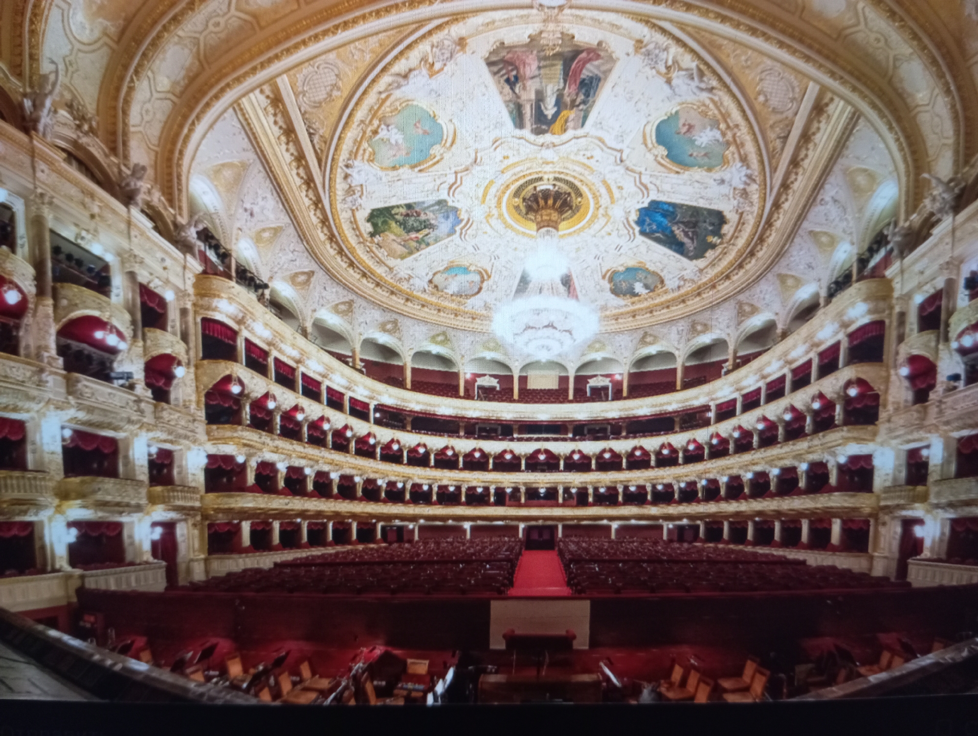 The image depicts the interior of the Odesa Opera and Ballet Theater in Odesa, Ukraine. The theater is empty. It shows the ornate, multi-tiered seating and balconies, the large chandelier hanging from the painted dome ceiling. The decor is primarily white and gold, with red velvet seats and curtains. The stage is visible at the bottom of the image. The room is brightly illuminated.