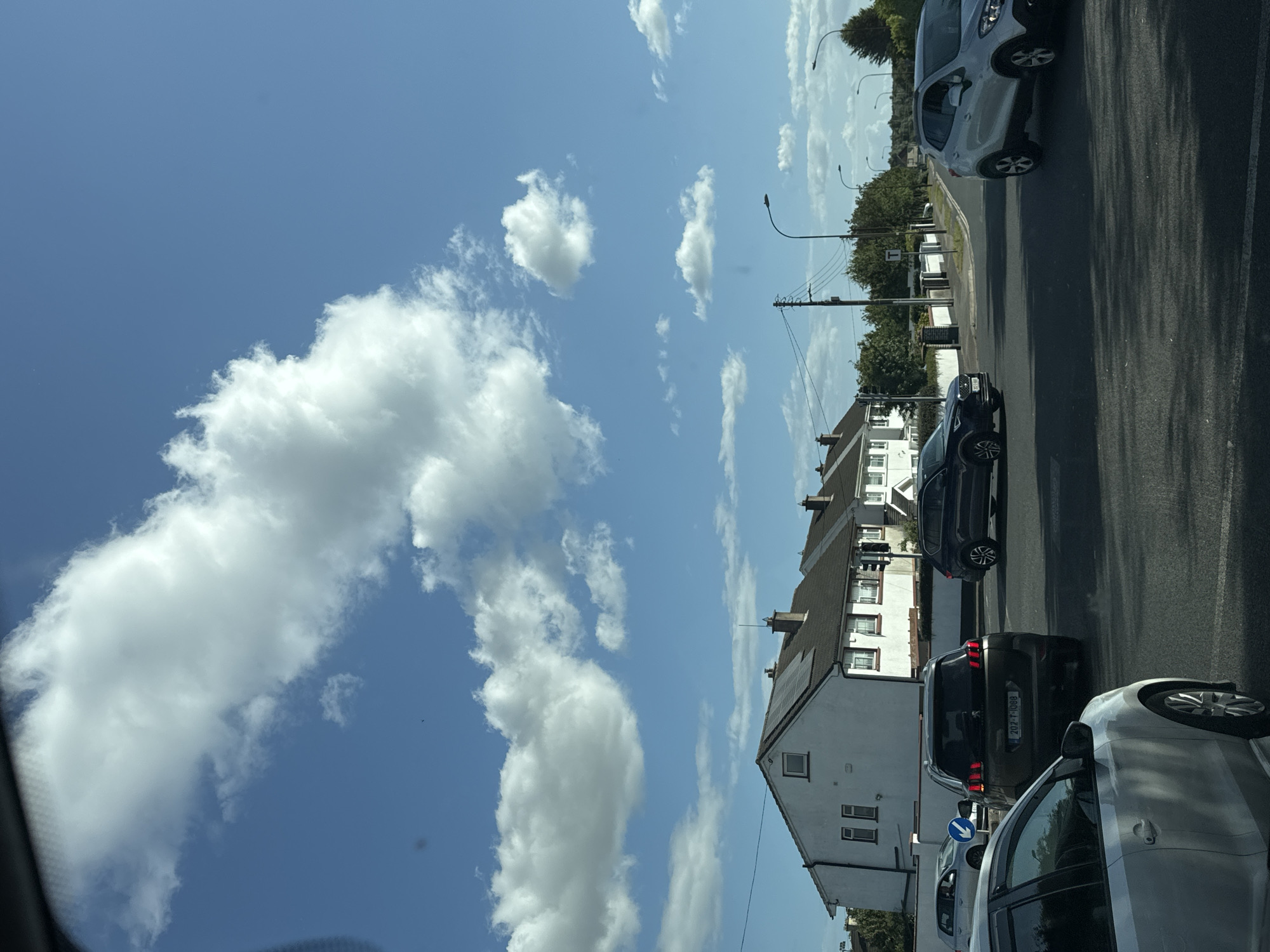 This is a ground-level, eye-level, medium shot of a suburban intersection in Waterford, Ireland, on a clear, sunny day with scattered white clouds. The foreground shows the asphalt road surface with lane markings and shadows cast by unseen objects. Several cars are visible at the intersection: a silver car in the immediate foreground, a dark-colored SUV to its left, a dark-colored Peugeot SUV in the middle lane with its rear facing the camera, and a silver SUV to the right. In the middle ground, rows of white, two-story houses with dark roofs line the street, some featuring solar panels. Streetlights and utility poles are also present. The background consists of more trees and houses, with a bright blue sky overhead. The overall scene depicts a typical suburban traffic junction under daylight conditions.