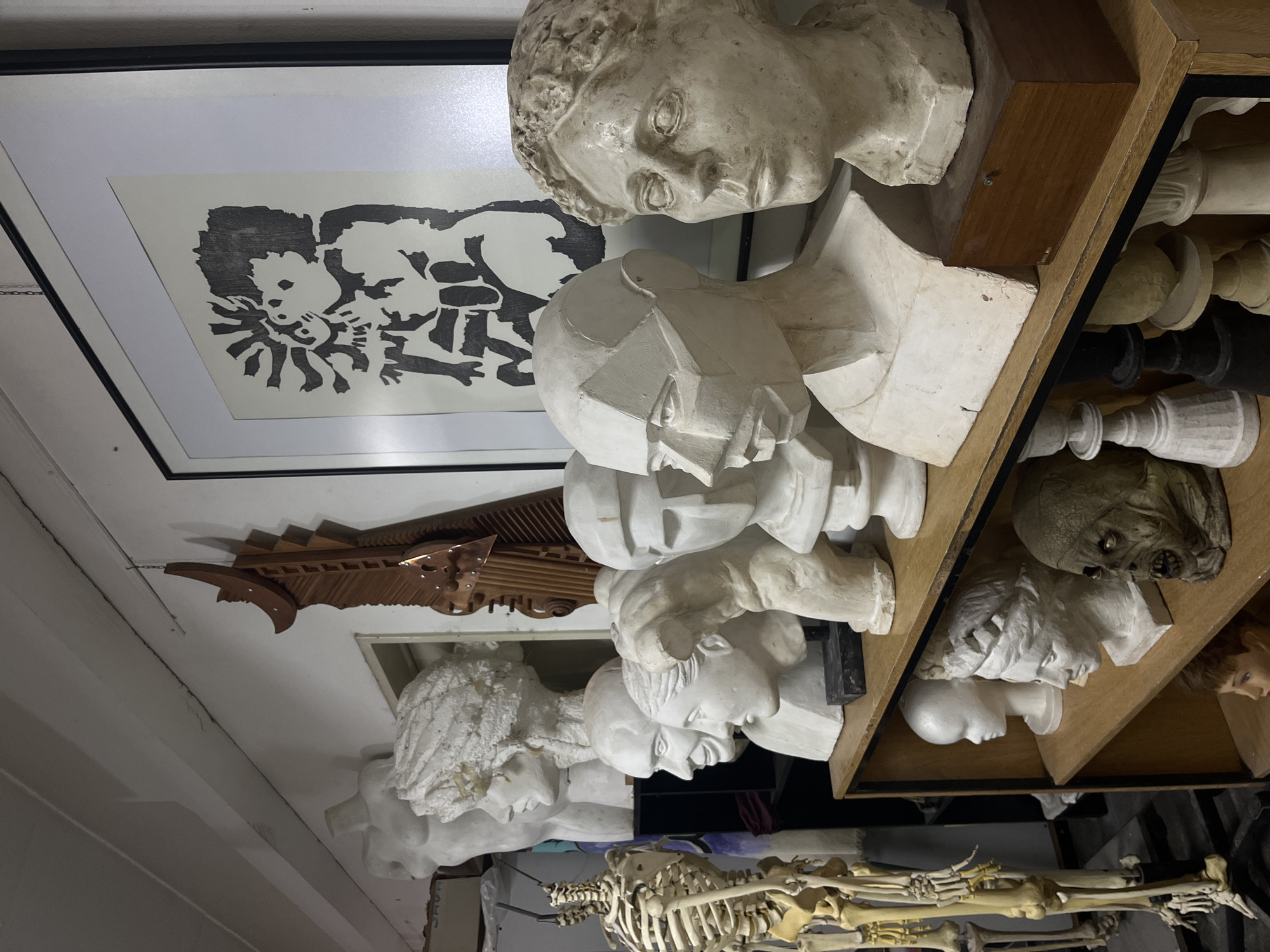 This media file captures an indoor scene, likely an artist's studio or a space dedicated to anatomical and artistic study, located in Buenos Aires, Argentina. The room is filled with a diverse collection of sculptures, models, and artwork, giving it a somewhat cluttered yet organized appearance.
On the left, a large, framed black and white print hangs on a light-colored wall. The artwork features bold, stylized figures with dark outlines and shapes against a white background, suggesting a linocut or woodcut style. Below the framed print, a full human skeleton model is visible, lying horizontally on what appears to be the floor.
To the right, a multi-tiered wooden shelving unit extends vertically, packed with numerous white and off-white plaster busts and head sculptures. The busts display a variety of styles, ranging from classical and realistic depictions with detailed features and hair textures to more abstract, geometric, and cubist-inspired forms. Among these heads, several large, ornamental chess pieces, both white and black, are arranged on the shelves. One shelf also holds a white foam mannequin head and a distressed, possibly aged, mask-like head sculpture. From beneath the shelves, another human-like doll or mannequin head can be seen peeking out.
In the mid-ground, above some of the lower busts, an intricately carved wooden sculpture, possibly a decorative architectural element or a part of a larger mechanical art piece, is mounted on the wall. The overall scene is static, showcasing a collection of items rather than any active event. No specific text is clearly legible within the image.