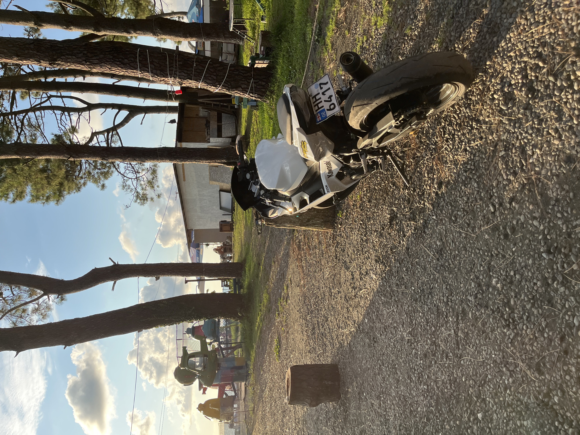The image depicts a scene in Ureki, Georgia, near the Black Sea coast. A white and black Kawasaki motorcycle is parked on a gravel surface in the foreground, leaning on its kickstand. The motorcycle has a license plate reading "HH 6417 GE". Behind the motorcycle, several tall pine trees are prominent. In the mid-ground, there is a white building with a wooden extension, and further back, a few amusement park rides, possibly for children, are visible. People are seated at tables in the background, and the sea is visible beyond the trees and rides. The lighting suggests it is late afternoon, with long shadows cast across the ground. The environment appears to be a recreational area, possibly a park or campground adjacent to a beach.
