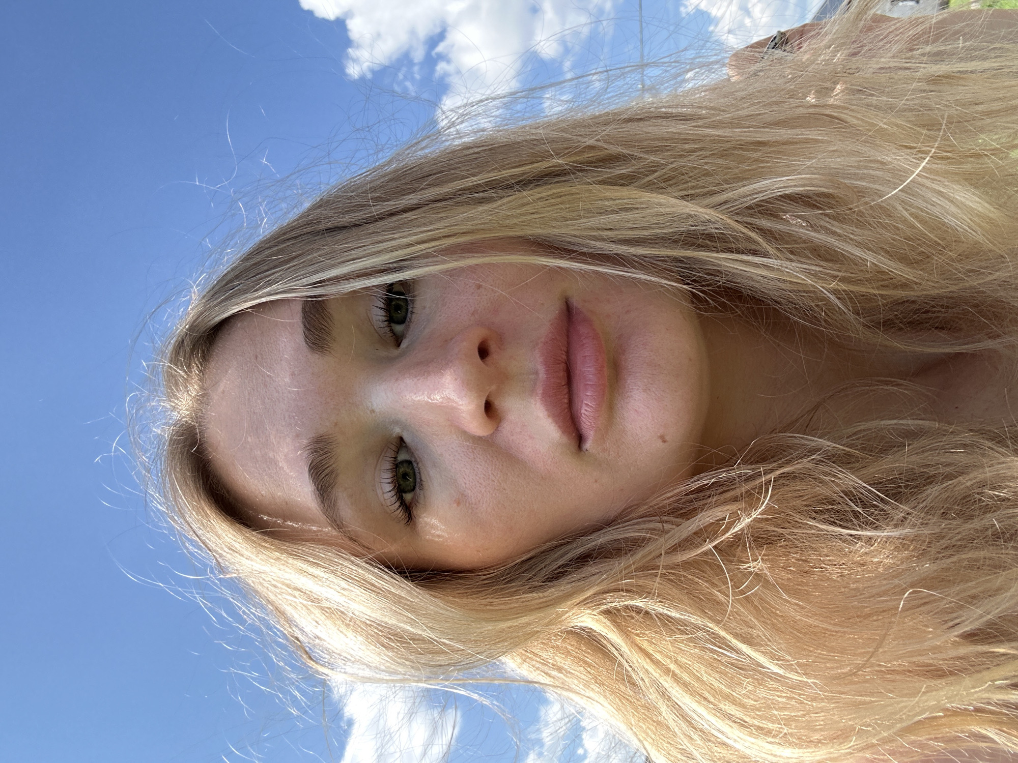 A young woman with blonde hair and green eyes looks directly at the camera, her face filling most of the frame. She has fair skin with freckles and appears to be outdoors, with a bright blue sky and some white clouds visible behind her. Her hair is slightly windblown, framing her face. The lighting suggests it is daytime, and the overall scene is casual and natural. There are no identifiable landmarks that would suggest a specific location within the city of Mokino, Russia.