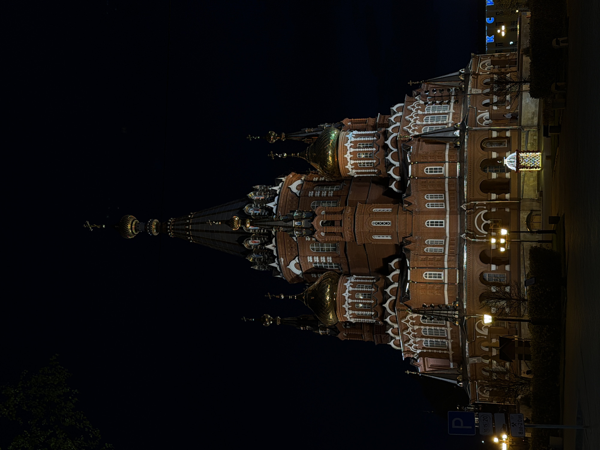 The image shows the National Museum of the Udmurt Republic in Izhevsk, Russia, at night. The museum, constructed of red brick with white detailing and topped with golden domes and a tall, spired tower, is illuminated by streetlights and interior lighting. The sky is dark, and sparse greenery is visible near the base of the building. Street signs and parking signs are on the lower right of the frame.