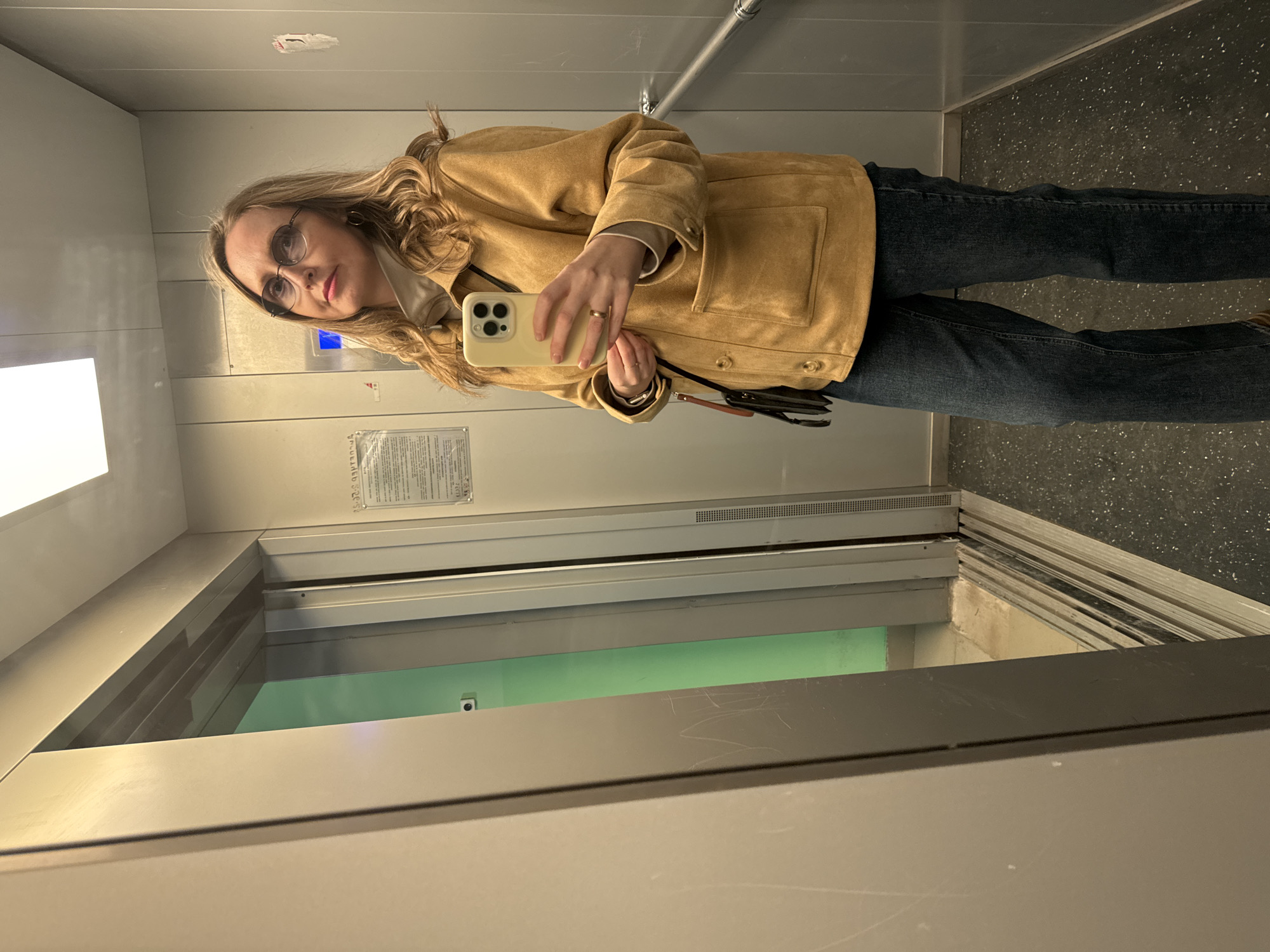 A woman is taking a selfie in what appears to be an elevator. She is wearing glasses, a tan jacket, and dark jeans. She is holding a phone in her right hand. The elevator has gray walls, a mirrored surface, and a dark speckled floor. A notice is posted on the elevator wall. The location is not explicitly identifiable as the city of Chaikovsky, Russia from the image alone.