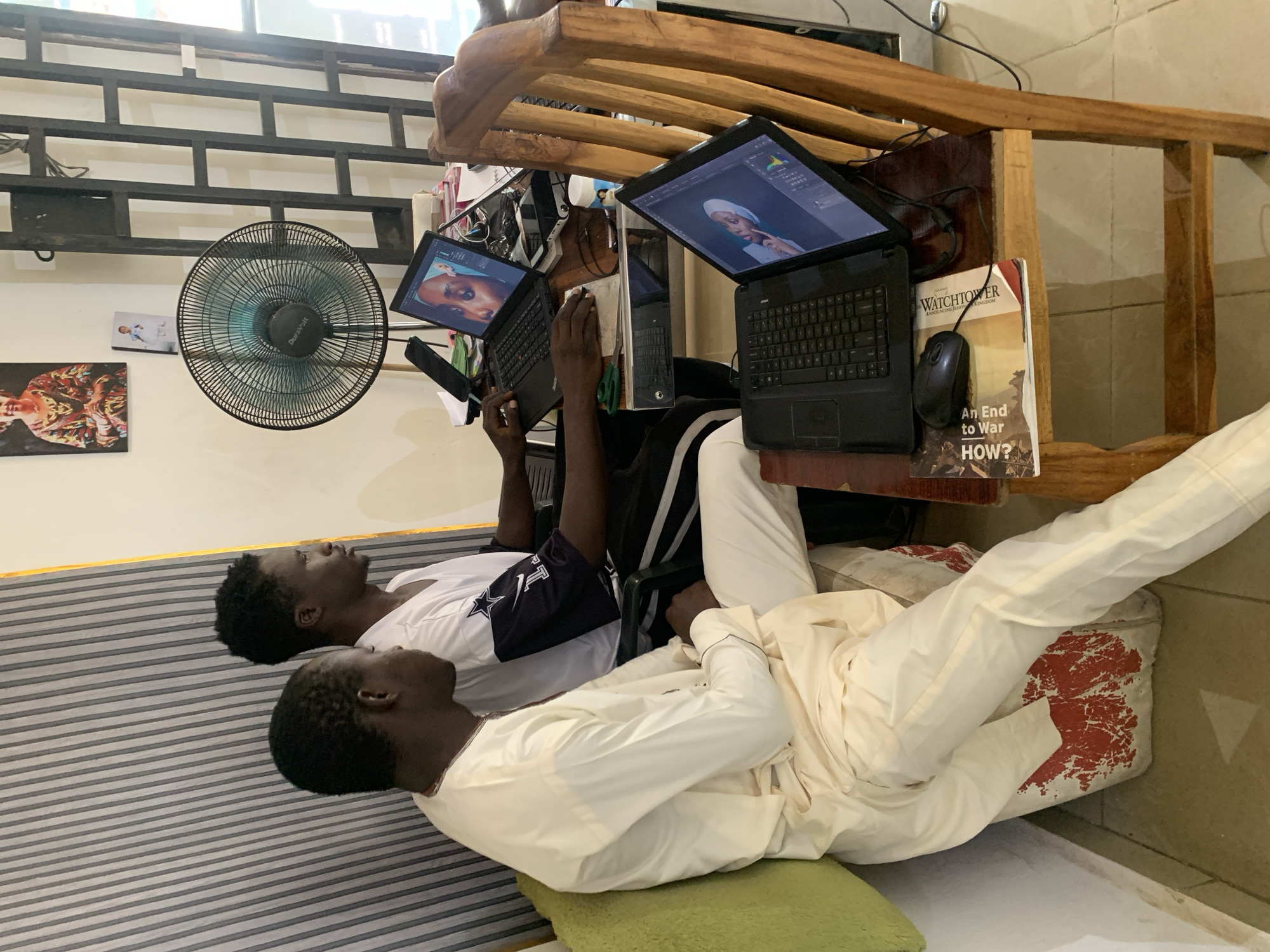 Two Black males are reclining on a horizontally striped surface in an indoor setting. One male wears a white shirt with a black-and-white logo and white pants, while the other is dressed in a full white outfit. They are positioned beneath a wooden table that supports two laptops with images displayed on their screens, a computer mouse, a book titled "WATCHTOWER," and a mobile phone. Above the table, an electric fan is mounted on the wall. Paintings are visible on the wall behind the fan. The context suggests that the location might be in Jos, Nigeria, although there is no explicit information to confirm this in the image itself.