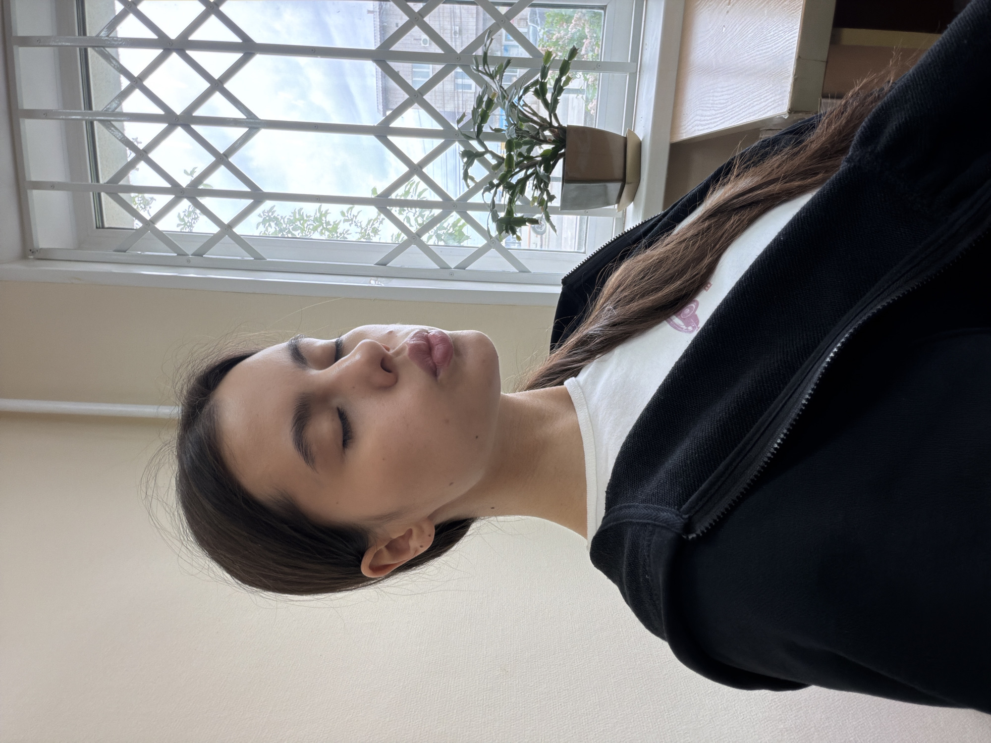 A young person, appearing to be female, is the main subject of the image, seen in a close-up, upward-angled view. She has long, dark brown hair, fair skin, and her eyes are closed. Her lips are slightly pursed, suggesting either a thoughtful expression, a "kiss face," or a moment of repose. She is wearing a white t-shirt visible under a black zip-up hoodie.
The setting is indoors, with a plain, light-colored wall filling the lower part of the frame. In the background, a large window is prominent, featuring a white, diamond-patterned metal security grille. Through the window, a bright, partially cloudy sky is visible, along with glimpses of distant buildings, indicating an urban or suburban environment in Chaikovsky, Russia. A small potted plant with green, segmented leaves rests on the windowsill, partially obscured by the person's head. The lighting is bright and natural, suggesting daytime. No discernible text is visible in the image.