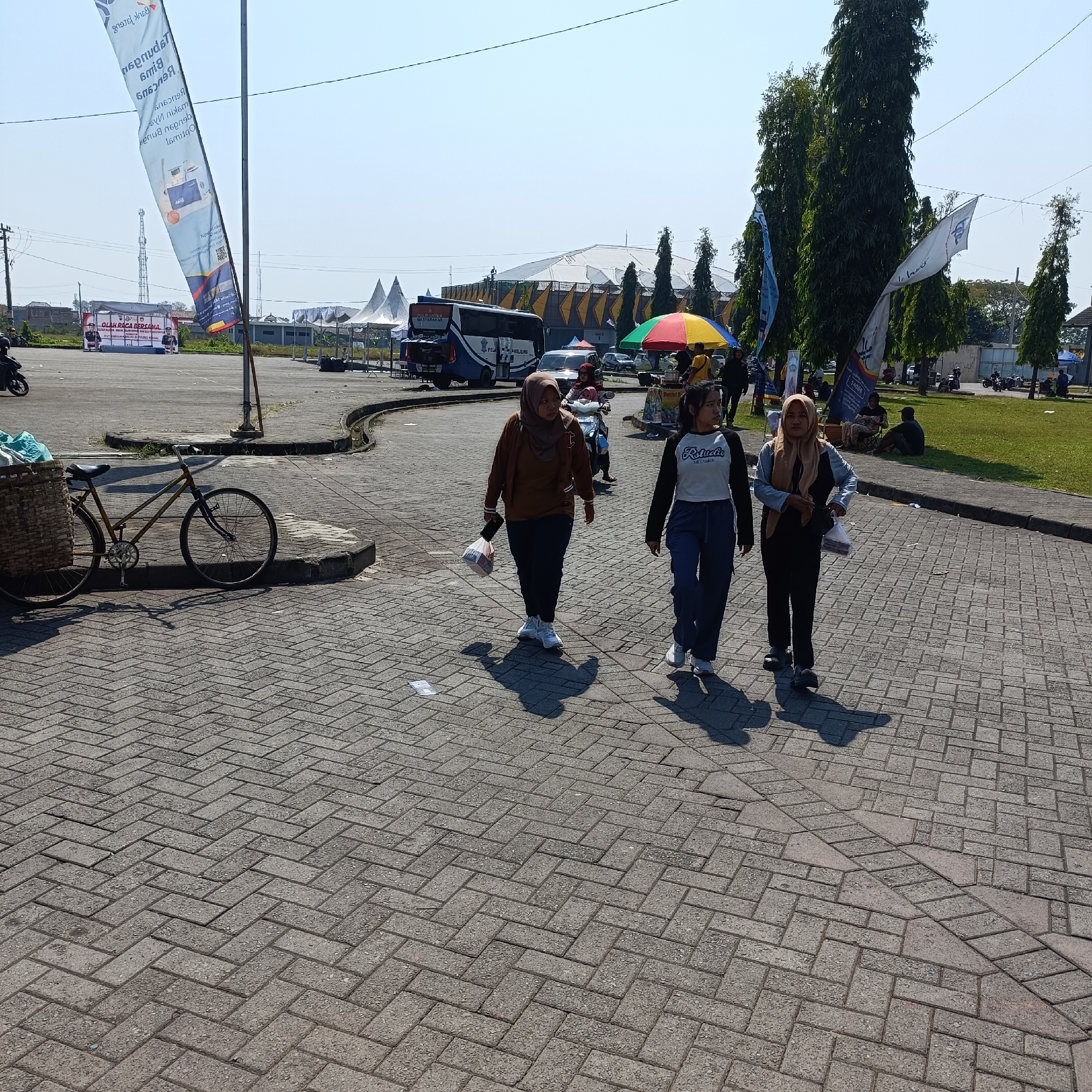 The image depicts an outdoor scene, likely in a public area with paved walkways and grassy areas. In the foreground, a bicycle with a basket is parked on the paving stones. Three women are walking across the paved area. The woman in the center is wearing a brown hijab and jacket, carrying a plastic bag. The woman to her left is wearing a black and white long-sleeved shirt and dark pants, while the woman to her right is wearing a light brown hijab and black pants.
In the background, there are tents, a bus, cars, and trees. Various flags are also visible, including a large one on the left with Indonesian text. The overall environment suggests an event or gathering is taking place. The presence of a sign with "KENDAL" visible on a flag suggests the location is Kendal, Indonesia. The weather appears sunny.