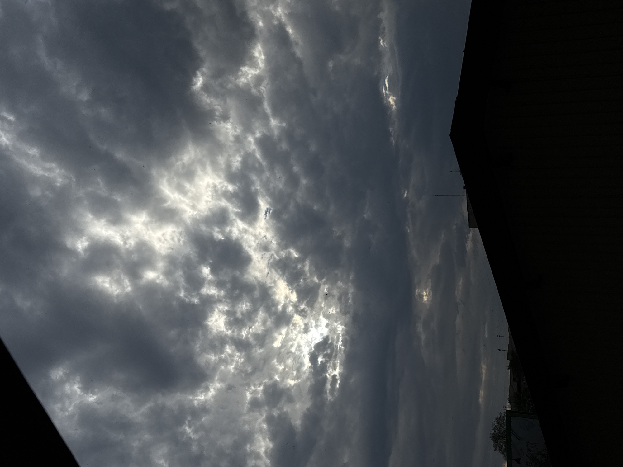 A dramatic, overcast sky looms over the city of Lobacheve, Ukraine. The clouds are dark and heavy, with patches of bright sunlight breaking through in the upper half of the image, creating a sense of impending rain or a storm. In the foreground, the dark silhouette of a building's roofline cuts across the lower portion of the frame, obscuring much of the cityscape below. Some indistinct structures and trees can be seen in the distance, barely visible beneath the moody sky. There are no visible people in the image.