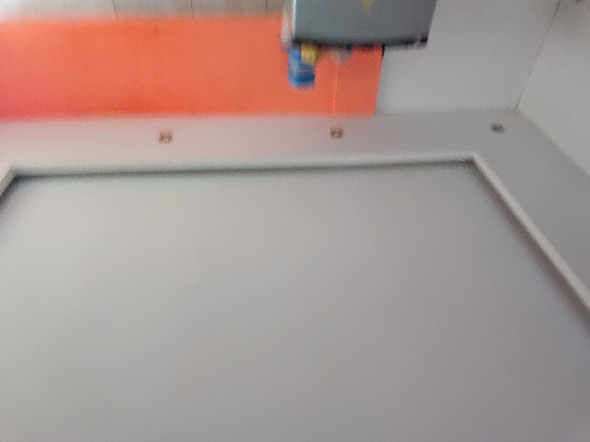 This media file presents a blurry, rotated indoor scene, likely a workshop or industrial facility, located in Santa Ana, Venezuela. The color palette is dominated by light grey, white, and a prominent orange.
A substantial, flat surface, primarily light grey, occupies the foreground and a significant portion of the midground. This surface appears to be a large work table, floor, or cutting mat, bordered by clean white edges or framing.
Above this grey surface, a large area of bright orange material, possibly a roll of vinyl, paper, or fabric, spans horizontally across the upper mid-section of the image. Directly above and seemingly interacting with this orange material is a rectangular machine with a grey housing. Attached to the underside of this machine are smaller components, including a distinct blue part and a yellow one. The machine's placement strongly suggests it is actively processing the orange material, perhaps as a large format printer, cutter, or laminator. The blur in the image is consistent with either camera shake or the motion of the machinery during operation.
Several small, dark, indistinct fixtures or fasteners are visible on the white border near the orange material in the mid-right section of the frame. The setting conveys a functional and utilitarian atmosphere. No people are visible, and there is no discernible text or branding. The time of day and weather cannot be determined as the scene is indoors with artificial lighting.