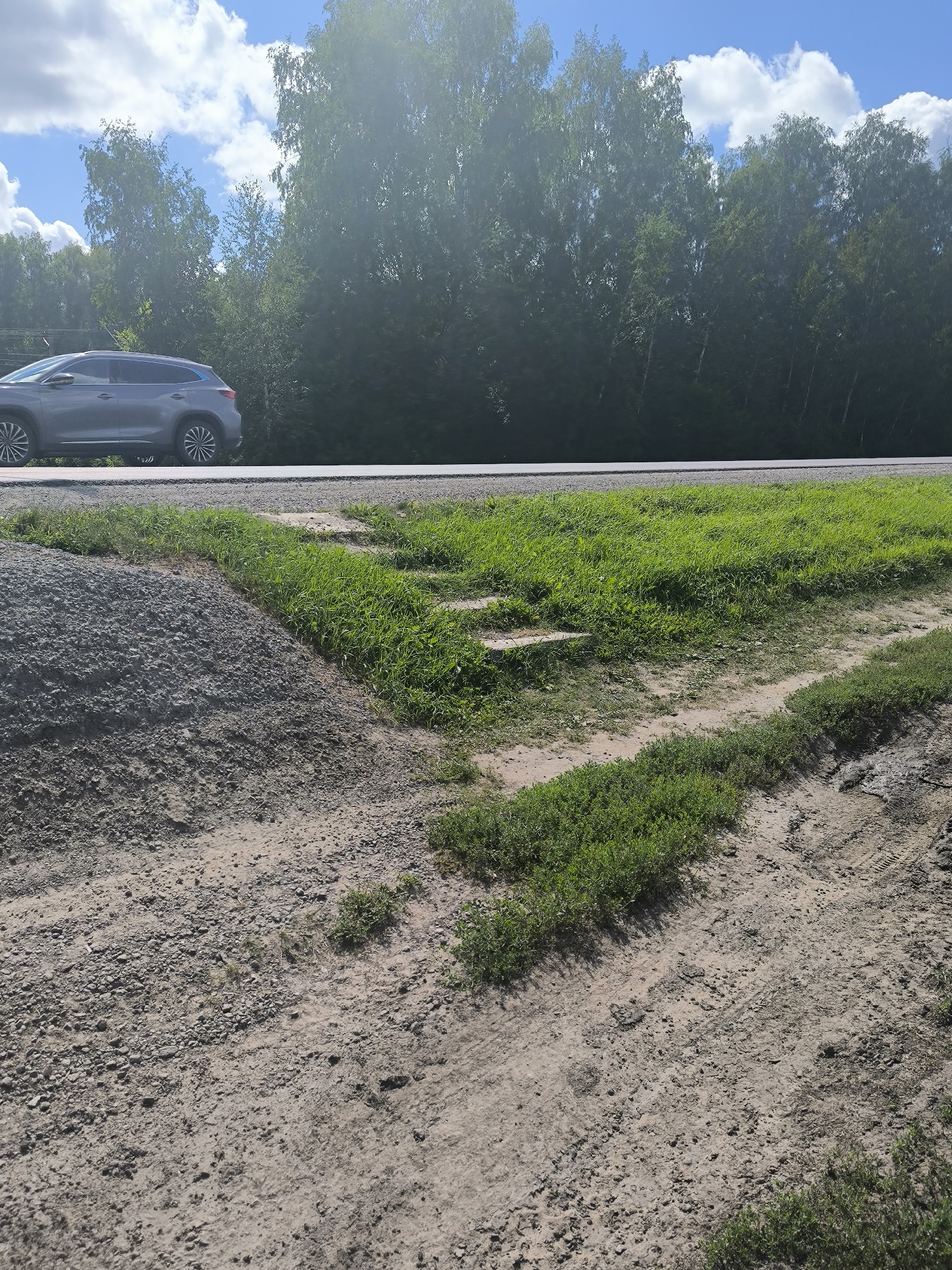 A gray SUV drives on a paved road bordered by a dense forest of deciduous trees under a bright blue sky with scattered white clouds. To the left of the road, a pile of gray gravel is visible. The foreground shows a dirt path with patches of green grass and tire tracks. The scene appears to be a rural or suburban area in the city of Maiskii, Russia.