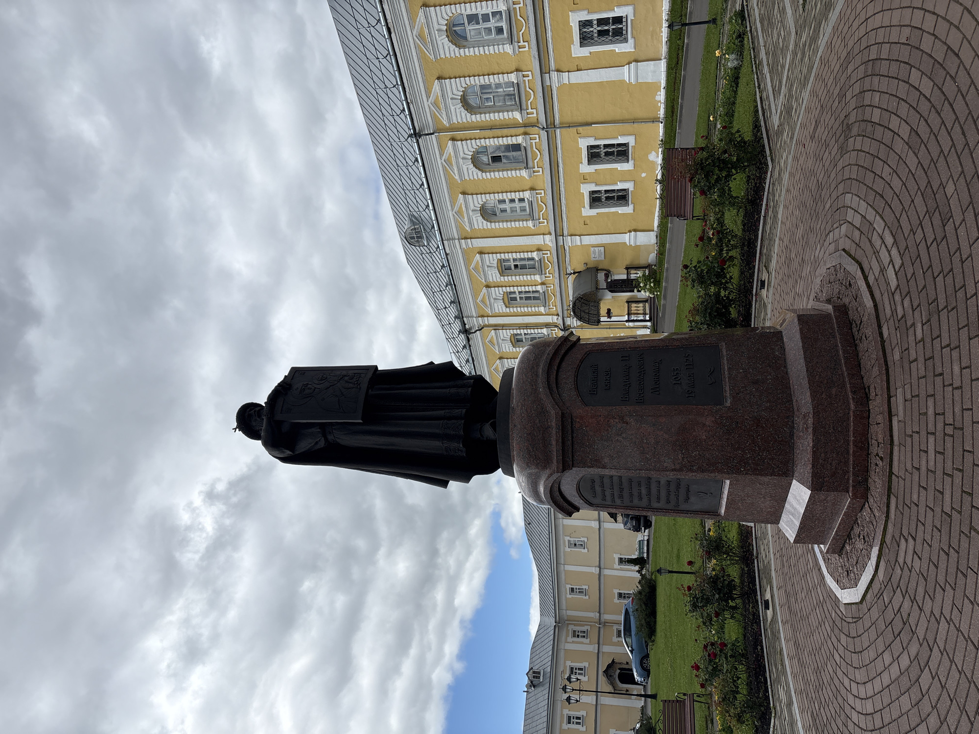 This is a medium shot, eye-level, full-body vertical photograph of a bronze statue of a robed figure holding a shield, set in a paved courtyard in front of a yellow building. The statue is positioned on a tall, multi-tiered pedestal made of dark red granite. The pedestal has inscriptions carved into its sides. The figure in the statue is wearing a long robe and a headdress, and is holding a shield with a relief design. The background features a long, two-story yellow building with white trim around the windows and roofline. The building has a grey, sloped roof with dormer windows and multiple arched and rectangular windows. To the left of the statue, a blue car is parked on a grassy area with rose bushes and benches. The sky is overcast with dramatic clouds. The paved area in the foreground is made of red brick, laid in a circular pattern around the statue's base. The location is identified as Sof'ino, Russia.