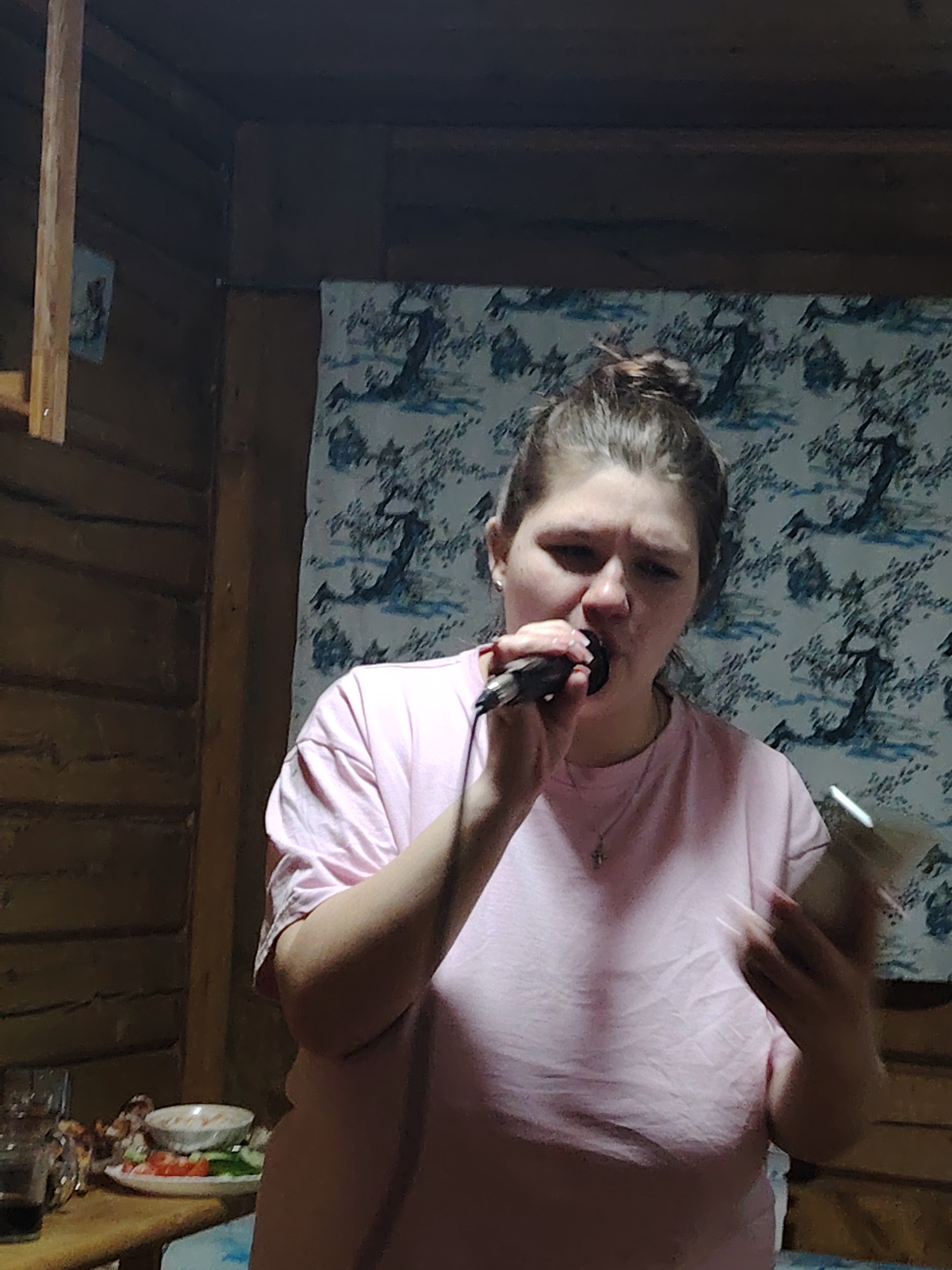 A woman is singing karaoke into a microphone. She is indoors, likely in a wooden house or cabin, as evidenced by the wooden wall and ceiling structure. Behind her is a patterned wall hanging. A table with food and drinks is visible in the foreground. The location is Malyi Bukor, Russia, but there are no details within the image to confirm that, only the query.