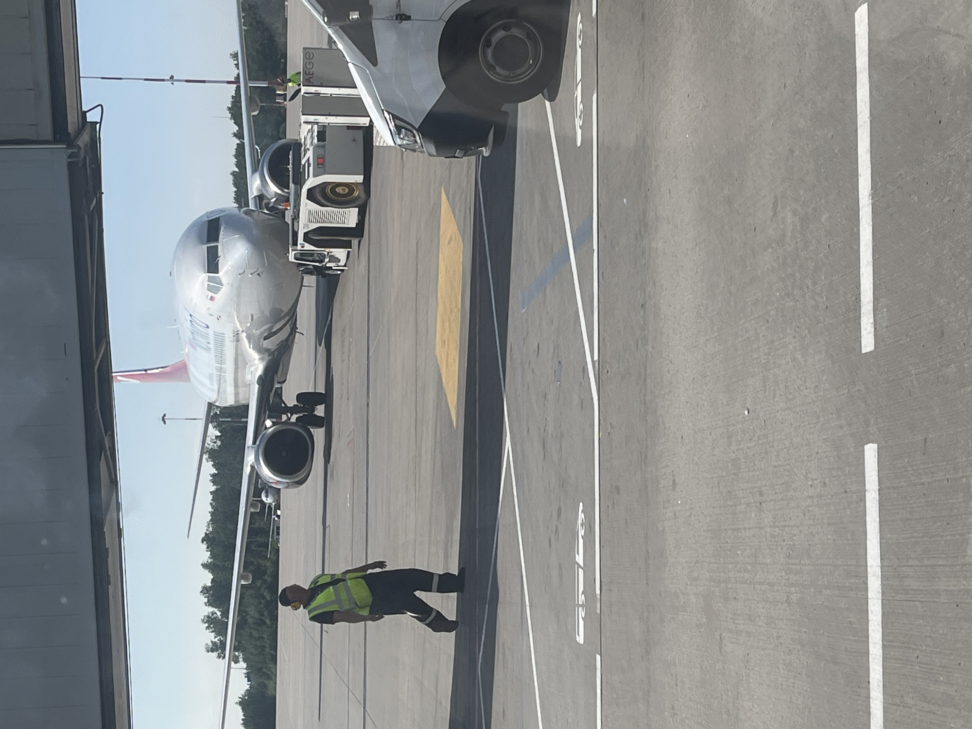 An airport scene in Saint Petersburg, Russia, is depicted. In the foreground, a man wearing a high-visibility yellow vest and dark pants walks on the tarmac. He is facing away from the camera, and his left arm is slightly bent. Behind him, a large commercial airplane, identified by a visible logo with Cyrillic script and possibly the airline "Smartwings," is parked. The aircraft is silver with a distinctive red and white tail. In front of the airplane's nose, a white pushback tractor is attached, with a person in a similar high-visibility vest standing near its rear. Further in the background, another aircraft can be seen parked. A white van is visible on the right side of the frame, partially obscuring the airplane. The ground is paved concrete with white lines marking parking or movement areas. The sky is clear and blue, indicating a sunny day. The scene appears to be an active apron area of an airport.