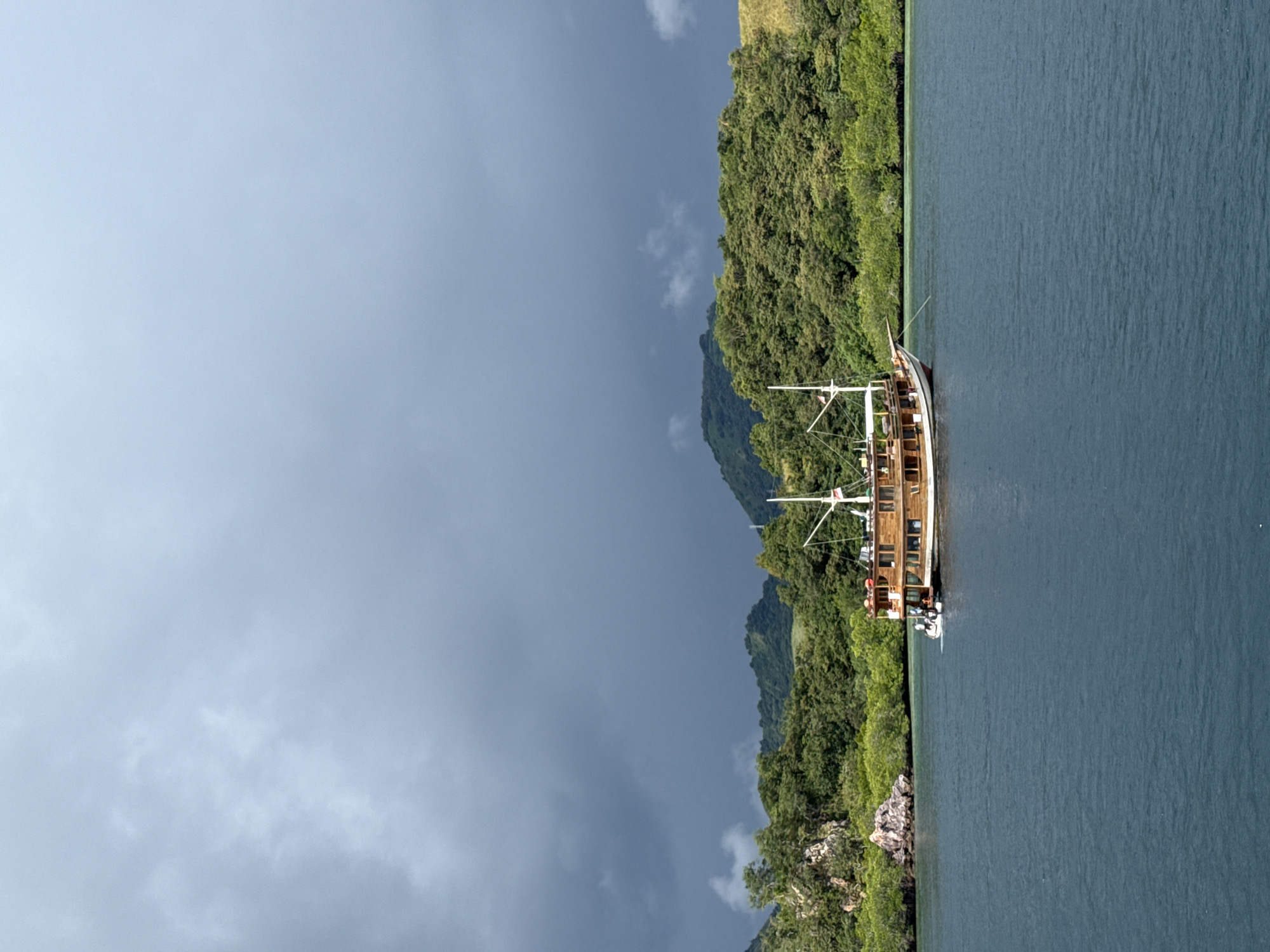 The image depicts a three-masted sailing vessel moored close to the shore of Rinca Village, Indonesia. The vessel, constructed largely of wood, is anchored in the water close to a densely forested shoreline. The terrain appears hilly, as lush green foliage climbs from the water's edge to the upper slopes. Above the verdant landscape, the sky is a muted gray, suggesting overcast conditions. The water itself is a deep blue, reflecting the color of the sky. There are people on the deck of the vessel. The scene gives the impression of a remote, natural setting.