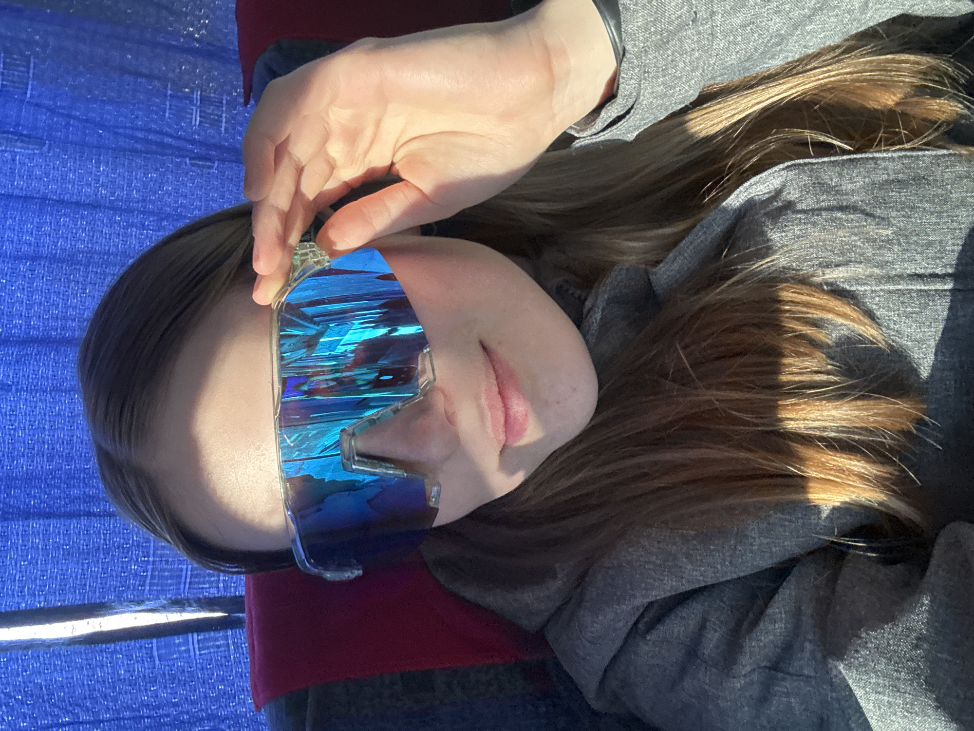 A person is wearing futuristic, reflective blue sunglasses, partially obscured by their right hand which is positioned to adjust the eyewear. The person has long blonde hair and is wearing a gray hooded sweatshirt. The background appears to be blue and possibly a seat. The location of Berezniki, Russia is not discernible from the image.