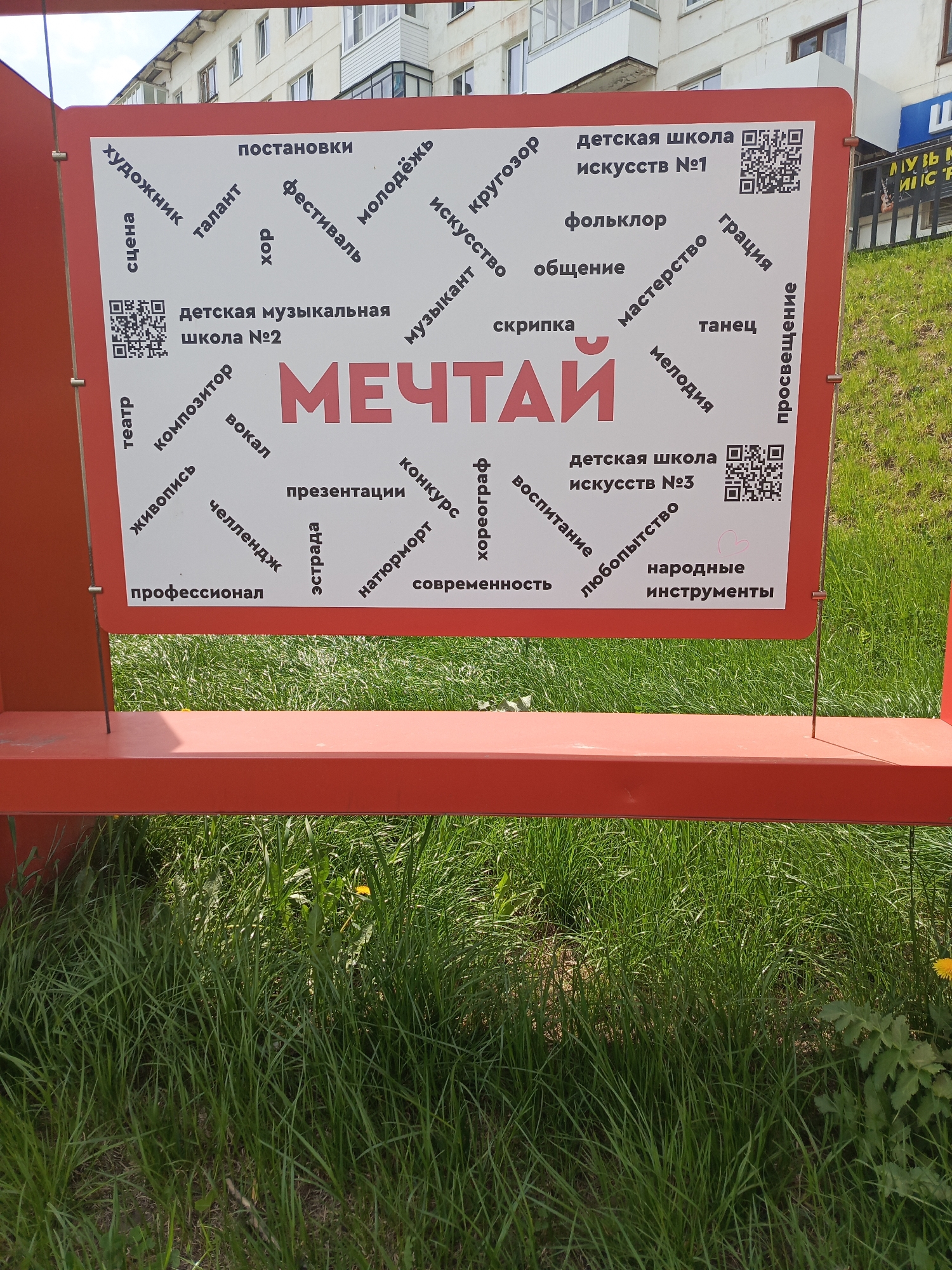 The image shows a red sign, partially obscured by grass, with the Russian word "МЕЧТАЙ" (roughly translated to "DREAM") printed in large red letters in the center. Around this central word are other smaller Russian words in black, seemingly associated with different artistic endeavors such as "художник" (artist), "сцена" (stage), "музыкант" (musician), "вокал" (vocal), and "танец" (dance). There are QR codes scattered near the phrases "детская школа искусств №1" (Children's School of Arts #1), "детская музыкальная школа №2" (Children's Music School #2), and "детская школа искусств №3" (Children's School of Arts #3). The sign is located in Chaikovsky, Russia. The immediate environment is characterized by a grassy area. In the background is a building.