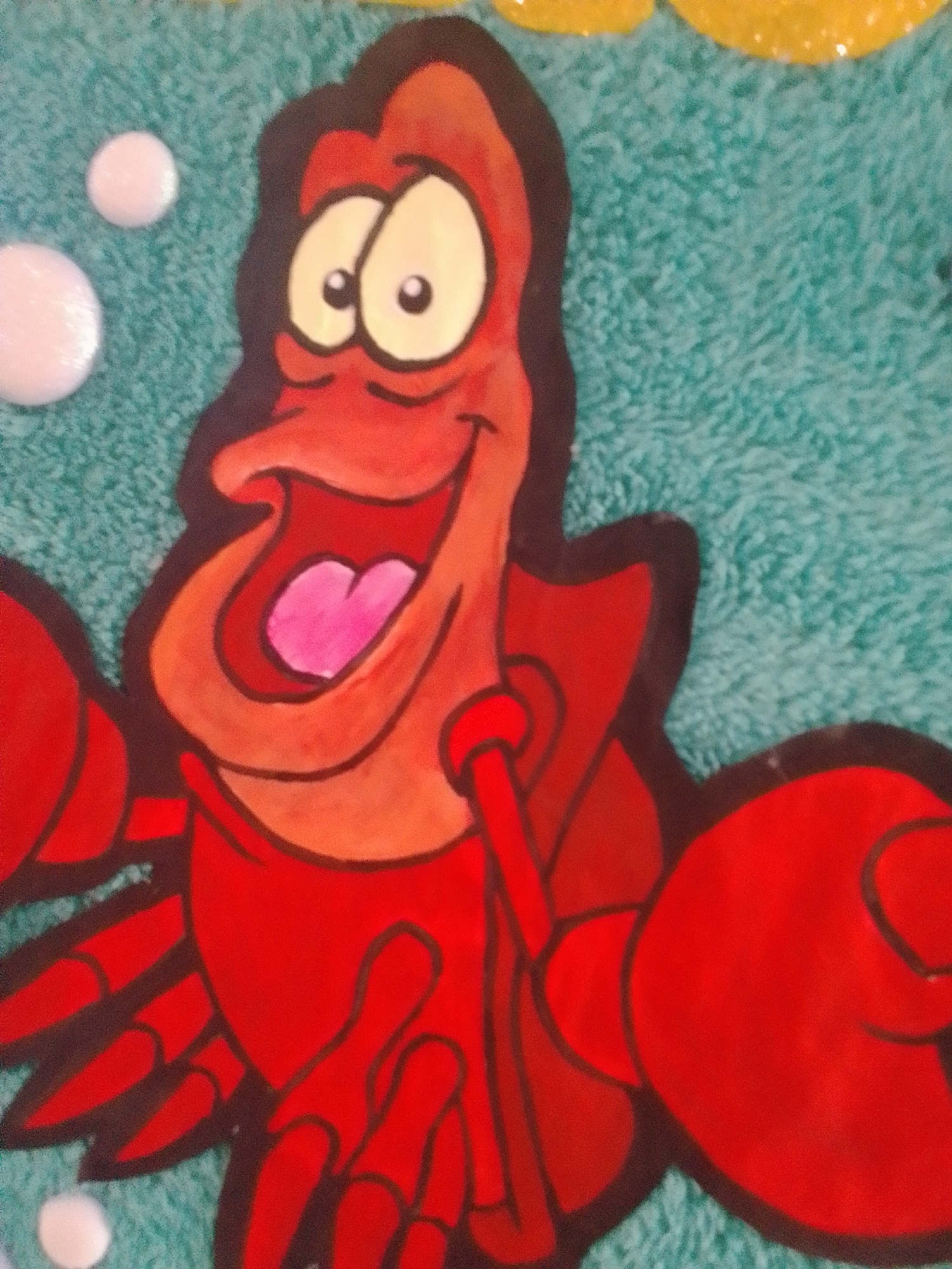 This media file shows a close-up of a cartoon crab character, Sebastian from Disney's "The Little Mermaid." The crab is depicted in his iconic red color, with large, expressive eyes and an open mouth revealing a pink tongue. He is drawn with bold black outlines, characteristic of animation. The background is a textured teal color, resembling water or perhaps a towel, with a few white circular shapes that could represent bubbles. In the upper right corner, there are hints of yellow, possibly indicating sunlight or a decorative element. The overall impression is a cheerful and lively character. The setting appears to be a surface with a soft, fuzzy texture. There is no visible text in the image. The image is likely a photograph of a decorated item, such as a towel, a wall mural, or an art piece.