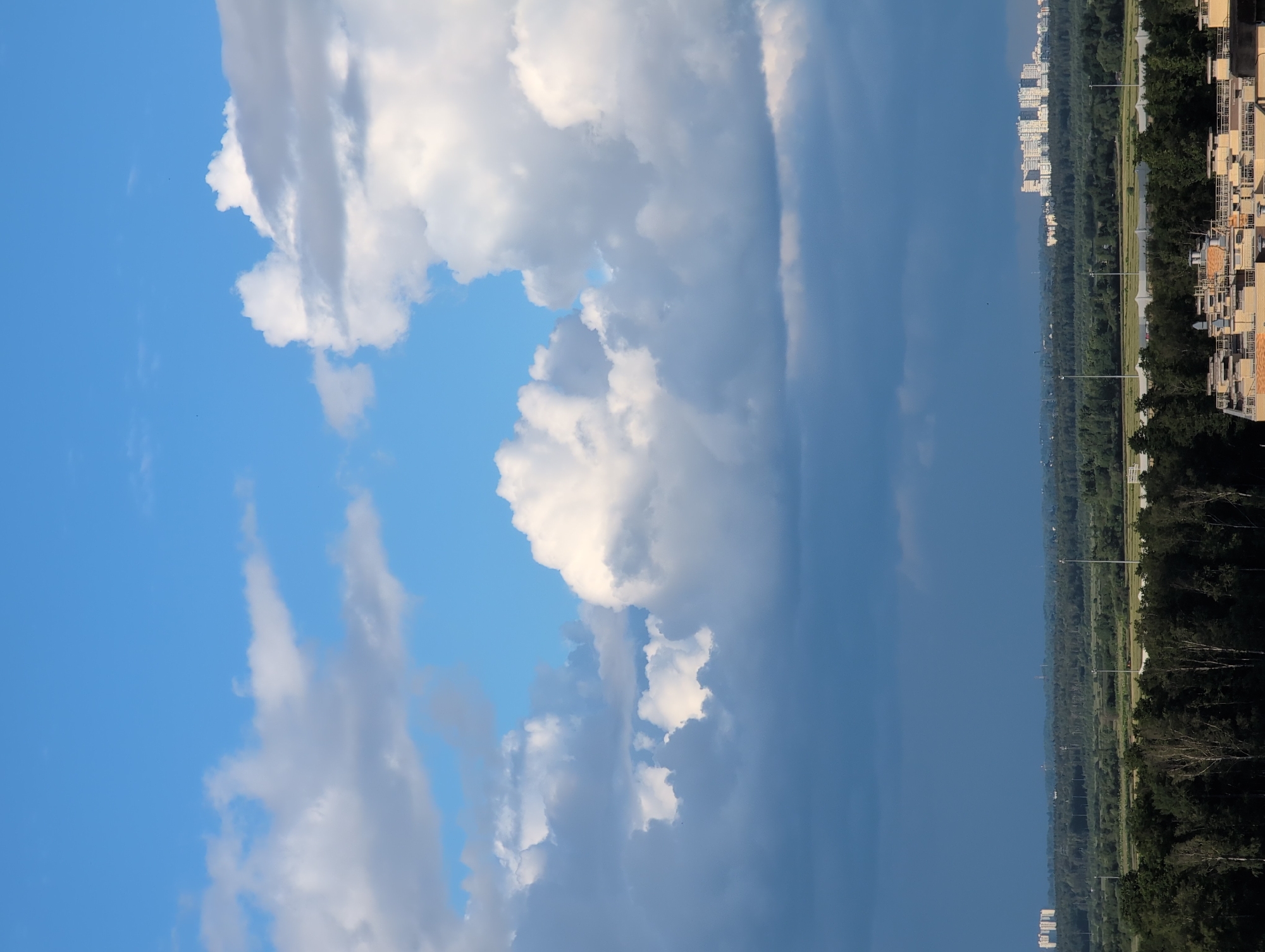 The image depicts a vibrant sky with scattered clouds. The sky transitions from a bright blue on the left to a darker, stormy gray on the right. Several white, fluffy clouds are prominent, catching the light.
Below the sky, a landscape unfolds. On the far right, the upper floors of apartment buildings are visible, suggesting an urban or suburban setting. These buildings are characterized by light-colored facades and numerous windows. Between the buildings and the horizon, a dense line of dark green trees forms a natural barrier. Beyond the trees, a long, straight road or pathway is visible, bordered by manicured green areas and punctuated by tall lampposts, hinting at a park or developed area. Further in the distance, more buildings and trees are discernible, fading into a hazy horizon.
The scene appears to be set during the daytime, with the lighting suggesting either morning or late afternoon. The presence of both bright blue sky and dark clouds indicates variable weather conditions, possibly a storm approaching or passing. The location context is Saint Petersburg, Russia, which aligns with the appearance of the architecture and the types of trees. There is no visible text in the image. The overall mood is one of dramatic natural beauty, with the stark contrast between the sky and the developed landscape.