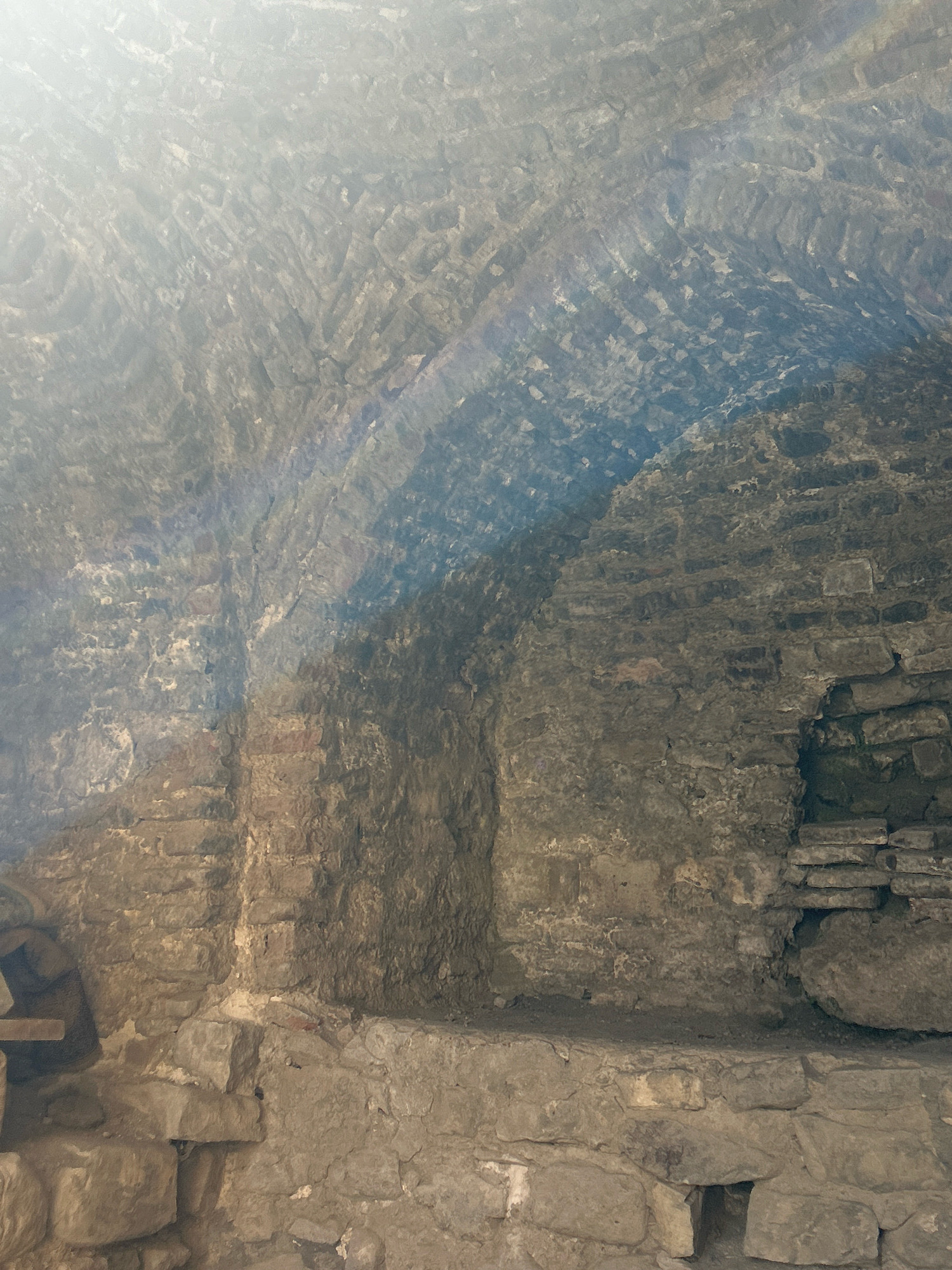 This image depicts an interior view of an ancient structure in Shalikiantkari, Georgia. The scene is characterized by rough-hewn stone and brickwork, forming arched ceilings and walls. A light beam, possibly from sunlight, illuminates the scene, casting shadows and highlighting the textures of the masonry. The construction appears old and possibly in a state of disrepair, with some stones dislodged and mortar showing wear. The environment is dimly lit and appears to be a historical site or ruin, with no people or specific objects of modern utility visible. The overall impression is one of antiquity and weathered resilience.