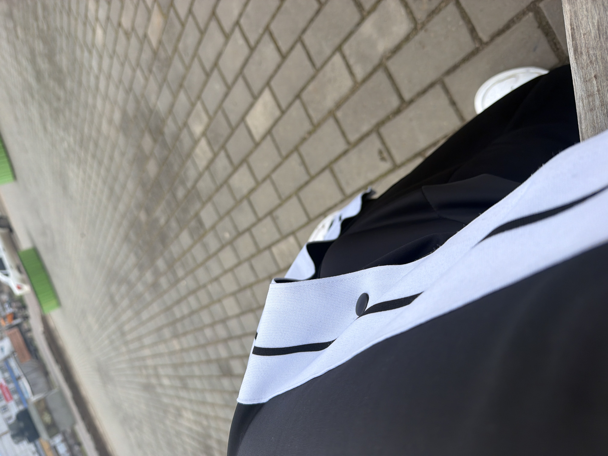 The image presents a first-person perspective looking down at a person wearing black clothing with white stripes and white shoes, seated on a bench beside a brick-paved walkway. The walkway leads to a street scene with buildings and vehicles, characteristic of an urban area in the city of Chaikovsky, Russia.
