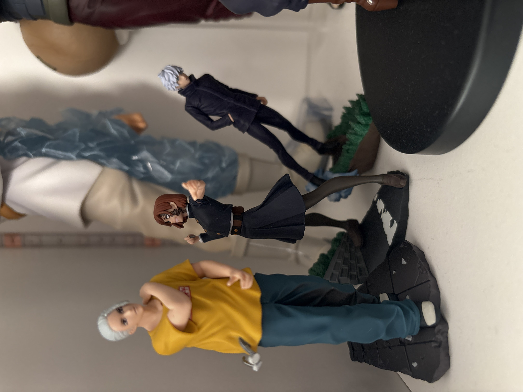 The image shows four anime figures displayed on a shelf. From bottom to top, they are: an older man with short white hair wearing a yellow t-shirt and blue pants, a woman in a black school uniform with brown hair, a slender male in a black suit with white hair, and a partially visible figure in red and tan clothing. The figures are all standing on black platforms with varying textures and details. The background is a plain, light-colored surface. The location is not discernible from the image.