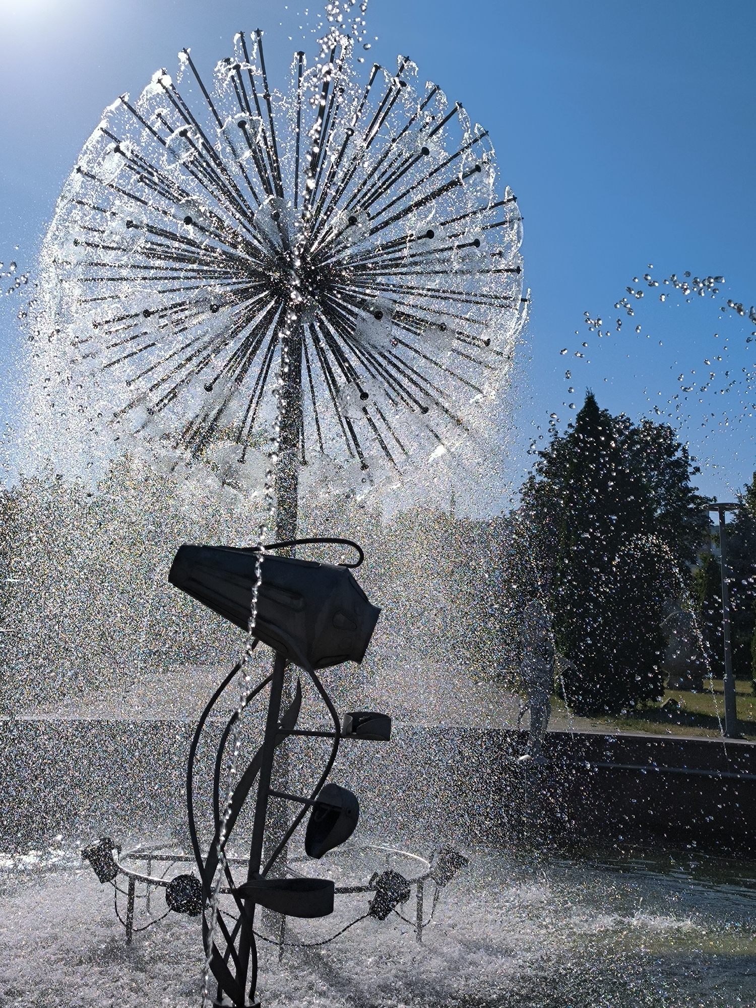This media file depicts a whimsical dandelion-shaped fountain with water spraying from numerous thin metal rods, creating a shimmering effect against a bright blue sky. The fountain has a central black structure with what appears to be a nozzle or spray head. The water cascades down, forming a misty curtain around the fountain's base. In the background, lush green trees are visible, suggesting an outdoor park or public space. The lighting indicates it's a sunny day, likely during daylight hours, as the sun's glare is noticeable on the water droplets. The overall scene evokes a sense of refreshment and playful artistry. The location is identified as Chișinău, Moldova. No people are visible in the frame, and no text is present.
