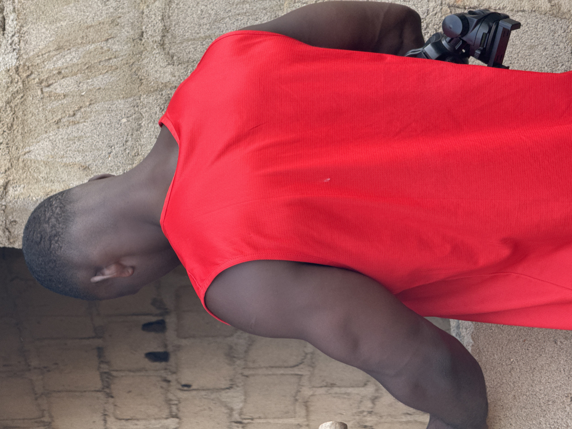 A person wearing a red sleeveless shirt is positioned with their back to the camera. They are standing next to a rough, sandy wall, and a brick wall with dark openings. A black camera stabilizer is attached to their right side, resting against the sandy wall. The scene appears to be outdoors, possibly in a construction or undeveloped area.