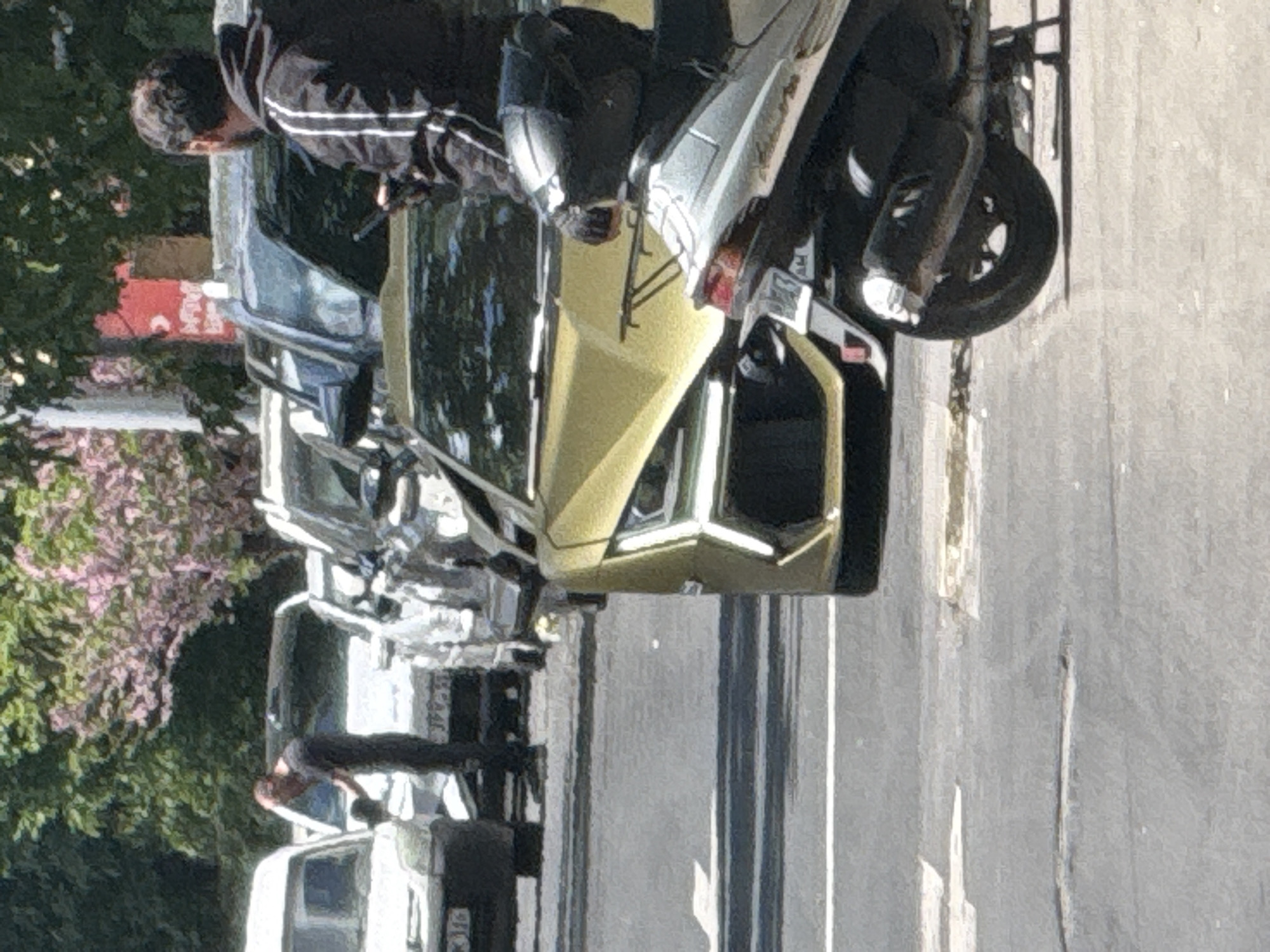 The image shows a street scene with a row of parked cars, a motorcycle and pedestrians. The cars range from older models to more recent designs. The motorcycle is parked on the side of the road, and a person wearing a black jacket with white stripes appears to be interacting with it. In the background, there are trees with pink blossoms, suggesting it could be spring. The street itself is paved and marked with painted white lines. The setting appears to be urban, likely Yerevan, Armenia, due to the style of vehicles and the architecture. There are multiple pedestrians visible, one in the middle of the frame.