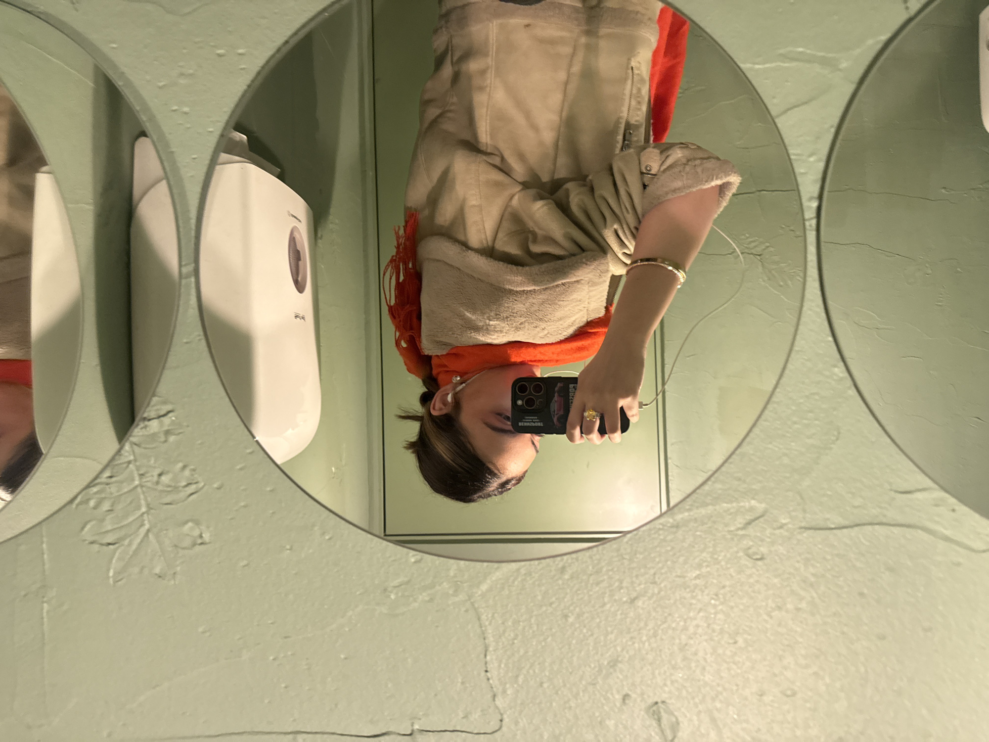 The image depicts a mirror selfie taken indoors in Yerevan, Armenia. A person is reflected from the chest up, wearing a light-colored (beige or tan) jacket with a shearling-like or fuzzy light-colored lining visible around the collar and cuffs, over a bright orange scarf with frayed ends. The person's dark hair is pulled back, and white wireless earbuds are visible in both ears. They hold a black smartphone in their right hand, which has a black case featuring a graphic resembling the rear of a sports car. A gold ring with a prominent yellow stone is on the person's right ring finger, and a gold-toned bracelet is on the right wrist. A white charging cable is plugged into the bottom of the smartphone. The surrounding walls, visible through and around the mirrors, are textured and painted a light sage green, showing minor cracks and faint decorative leaf patterns in some areas. The mirror arrangement consists of multiple overlapping or closely placed circular mirrors, creating a scalloped outline. To the right of the person's reflection, a white wall-mounted dispenser (likely for soap or paper towels) is partially visible.