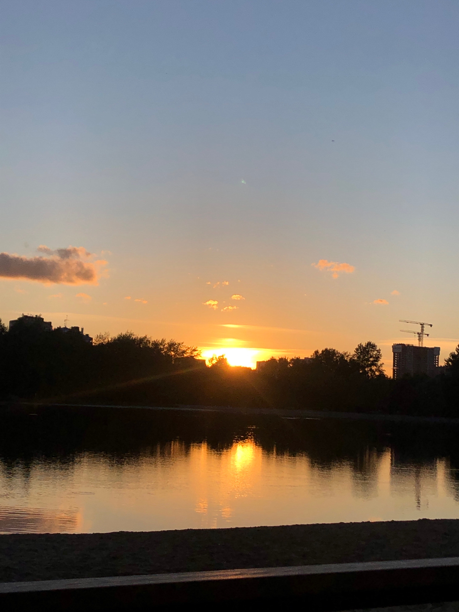 A serene sunset over a body of water, possibly a river or lake, is captured in this vertical outdoor shot. The sky transitions from a clear, pale blue at the top to a warm orange and yellow near the horizon, where the sun is setting. A few wispy clouds are scattered across the sky, tinged with the sunset's colors.
The foreground features a dark silhouette of trees and buildings along the water's edge. Among these are apartment buildings, with one prominent structure featuring construction cranes, suggesting ongoing development. The water itself reflects the vibrant colors of the sky, creating a shimmering, mirror-like surface. In the distance, the silhouette of more trees and structures can be discerned against the bright horizon. The bottom of the image shows a dark, sandy embankment, possibly a beach or park area.
The scene evokes a sense of peace and tranquility, typical of a sunset in an urban or suburban environment. Given the mention of Khimki, Russia, this location could be a park or waterfront area within the city, offering residents a place to enjoy the natural beauty of the sunset. The presence of construction indicates that Khimki is a developing city.