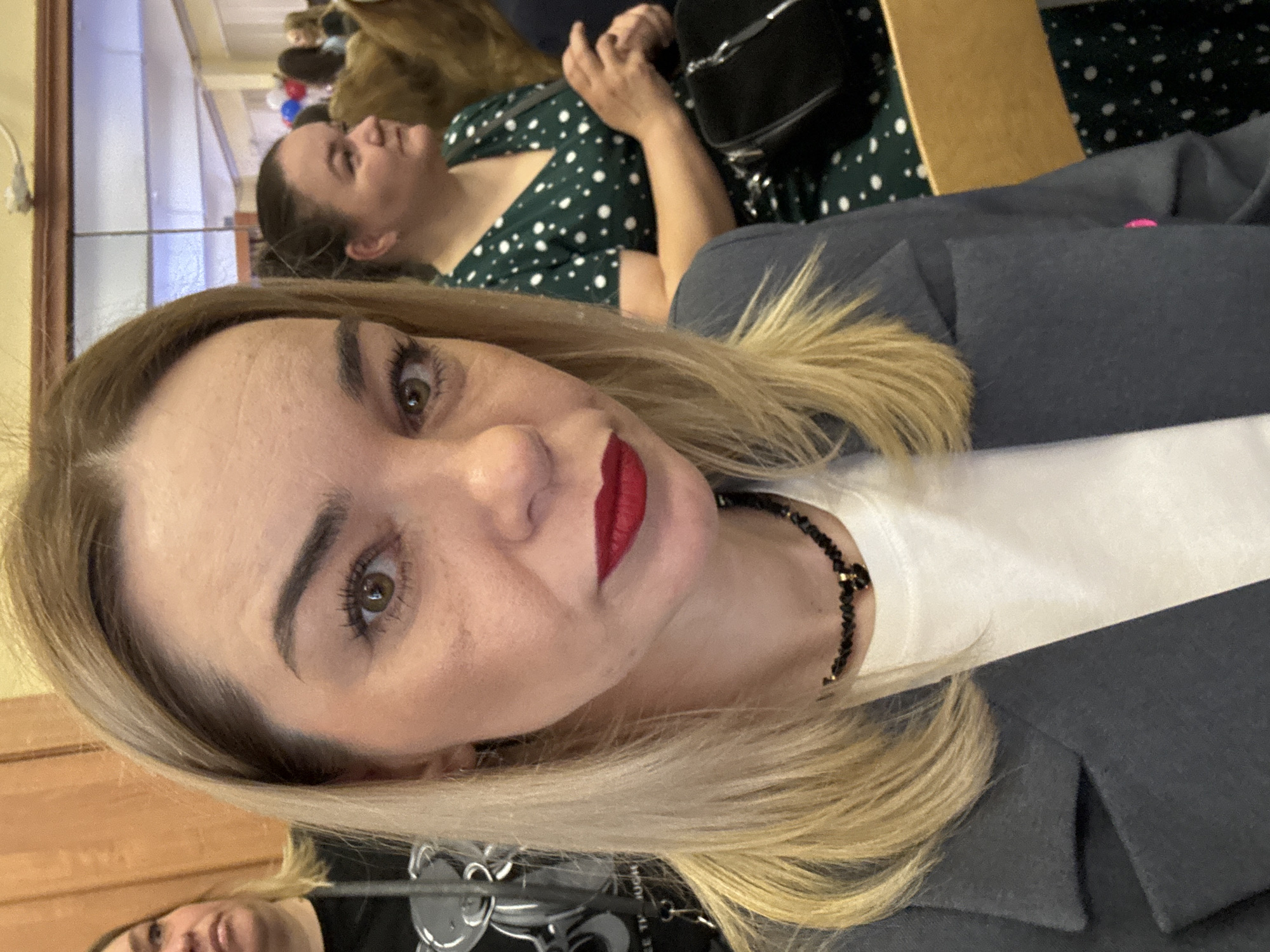 The image is a close-up selfie of a woman with blonde hair and red lipstick. She is wearing a white shirt and a dark gray blazer. A black beaded necklace is visible around her neck. Another woman with dark hair wearing a green dress with white polka dots sits behind her. A black purse is on the lap of the woman in green. The location is possibly indoors, as suggested by the lighting and the presence of other people in the background. The city of Murino, Russia, is not evident from the image.