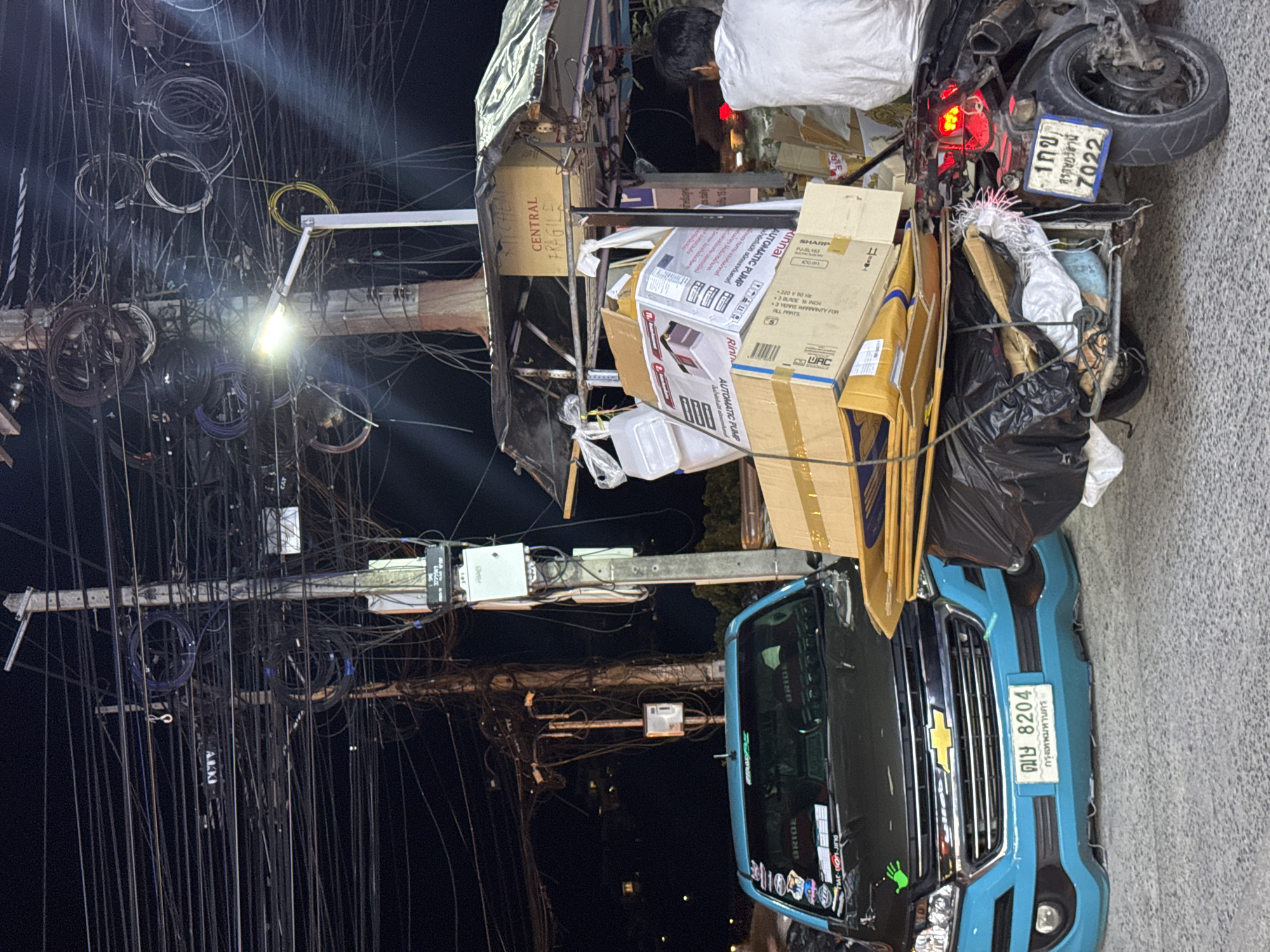 In an urban nighttime scene, a blue Chevrolet pickup truck with a black hood is parked on a paved road. The truck displays a license plate with Thai characters and the number "8204". To the right of the truck, a motorcycle with a sidecar is heavily laden with various items, including boxes, bags of trash, and what appears to be household appliances. A person wearing a white shirt is seated on the motorcycle, facing away from the camera. Overhead, a dense network of electrical wires is suspended from utility poles, illuminated by a streetlamp. A small vendor stall with a black tarp roof is situated behind the motorcycle, also displaying boxes. The overall environment suggests a busy street or market area.