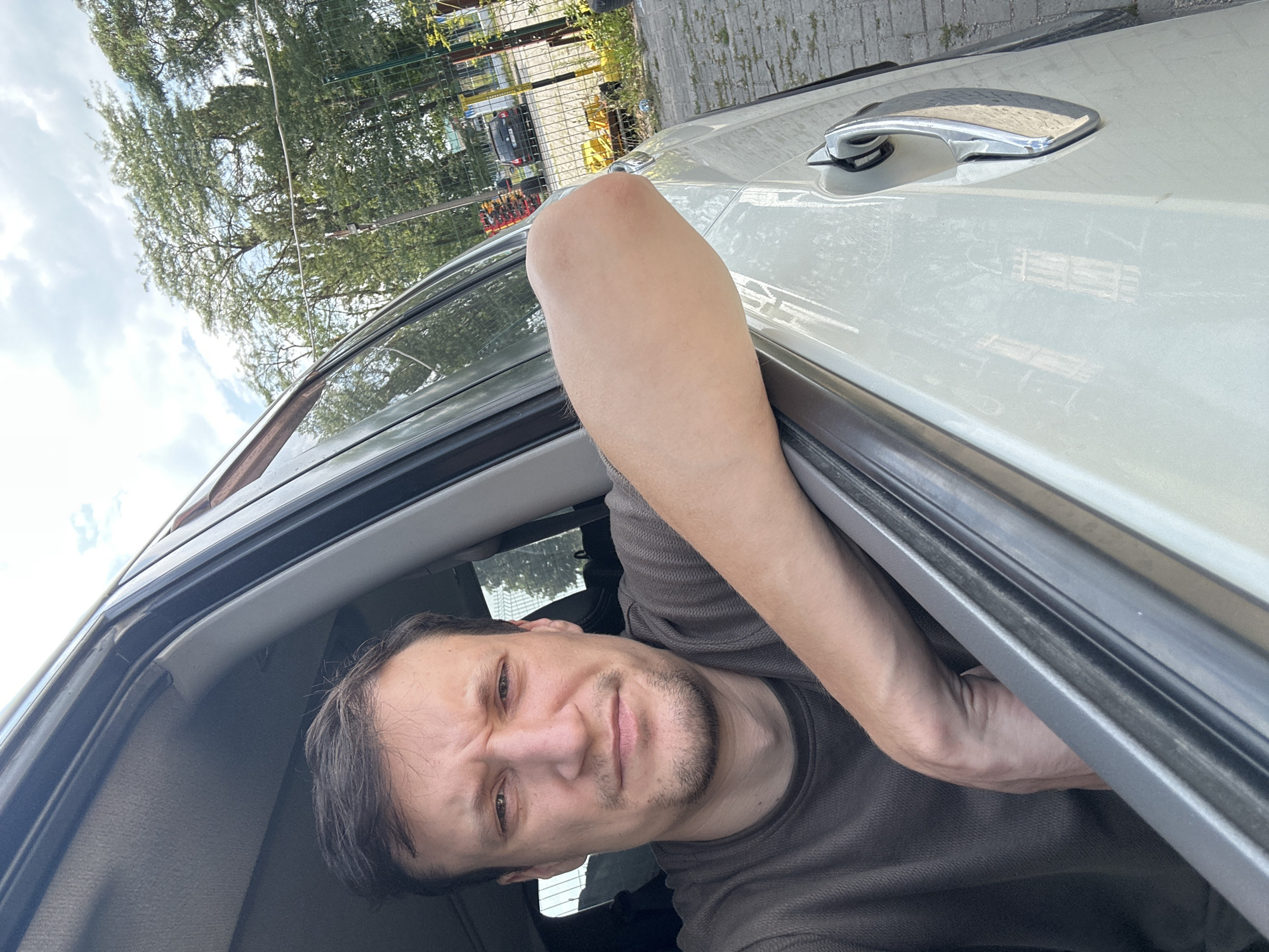 A selfie taken from inside a car shows a white man with short brown hair and a light beard. He is looking directly at the camera with a concerned expression, his brow furrowed. He is wearing a dark brown t-shirt. His right arm is resting on the open car door, with his elbow protruding slightly. The interior of the car is visible, including part of the roof lining and the car door frame. Outside the car, through the open window, trees and a fence are visible. A paved area, possibly a parking lot, is in the foreground. In the background, a dark-colored car is parked behind the fence. The sky is partly cloudy. The image is taken in Shatava, Ukraine.