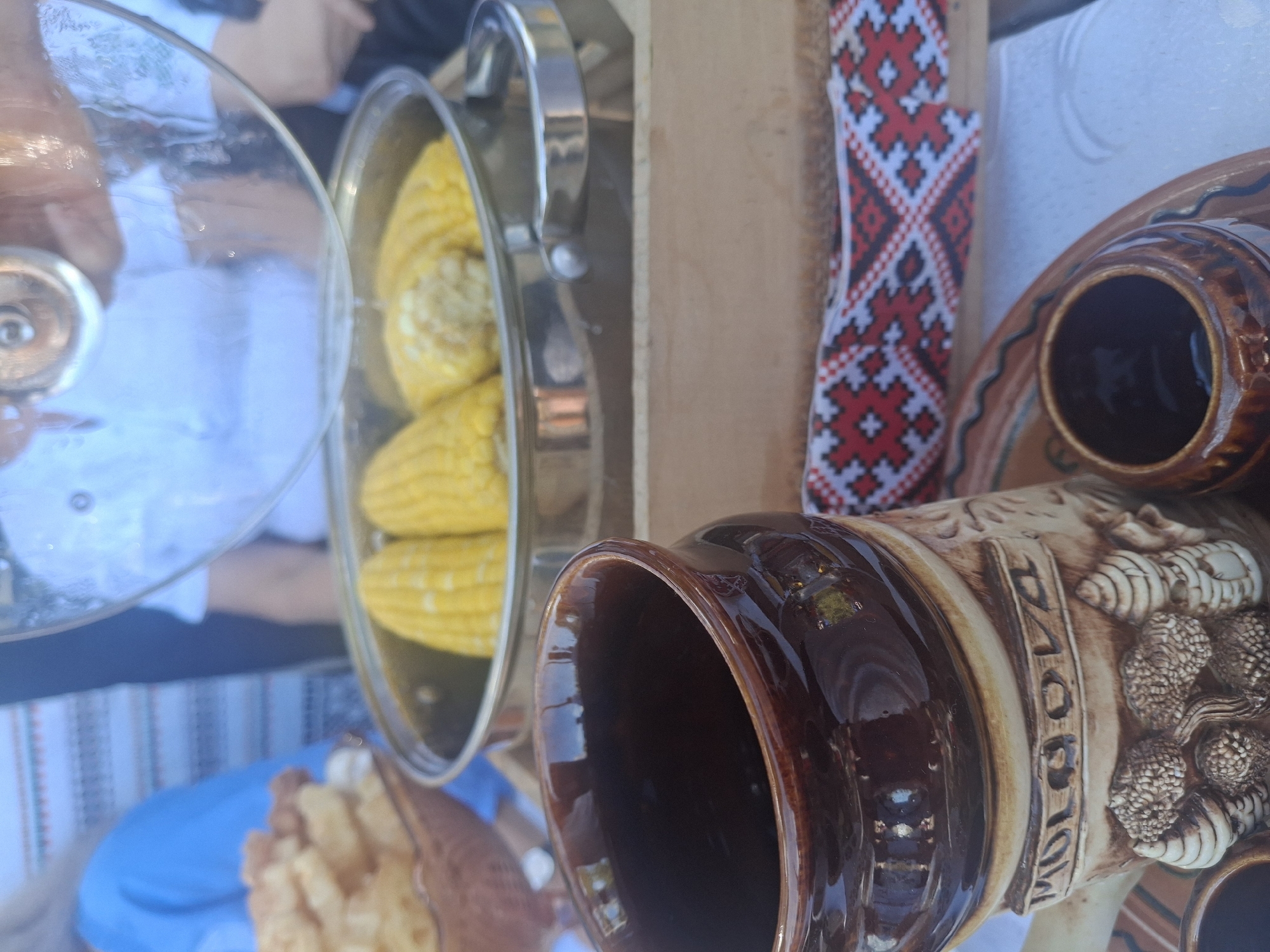 In Roghi, Moldova, a close-up shot focuses on a rustic table setting with traditional elements. A partially lifted glass lid reveals steaming ears of corn in a metal pot. To the left, a woven basket holds fried food, possibly chips or similar snacks. In the foreground, a decorative brown ceramic mug with embossed lettering and detailing sits prominently, suggesting it is a souvenir or traditional drinkware. A small brown ceramic cup and a decorative plate with intricate patterns are also visible. A red and white embroidered fabric band runs along the edge of a wooden serving tray. The background is blurred but suggests an outdoor setting, with hints of people and clothing.