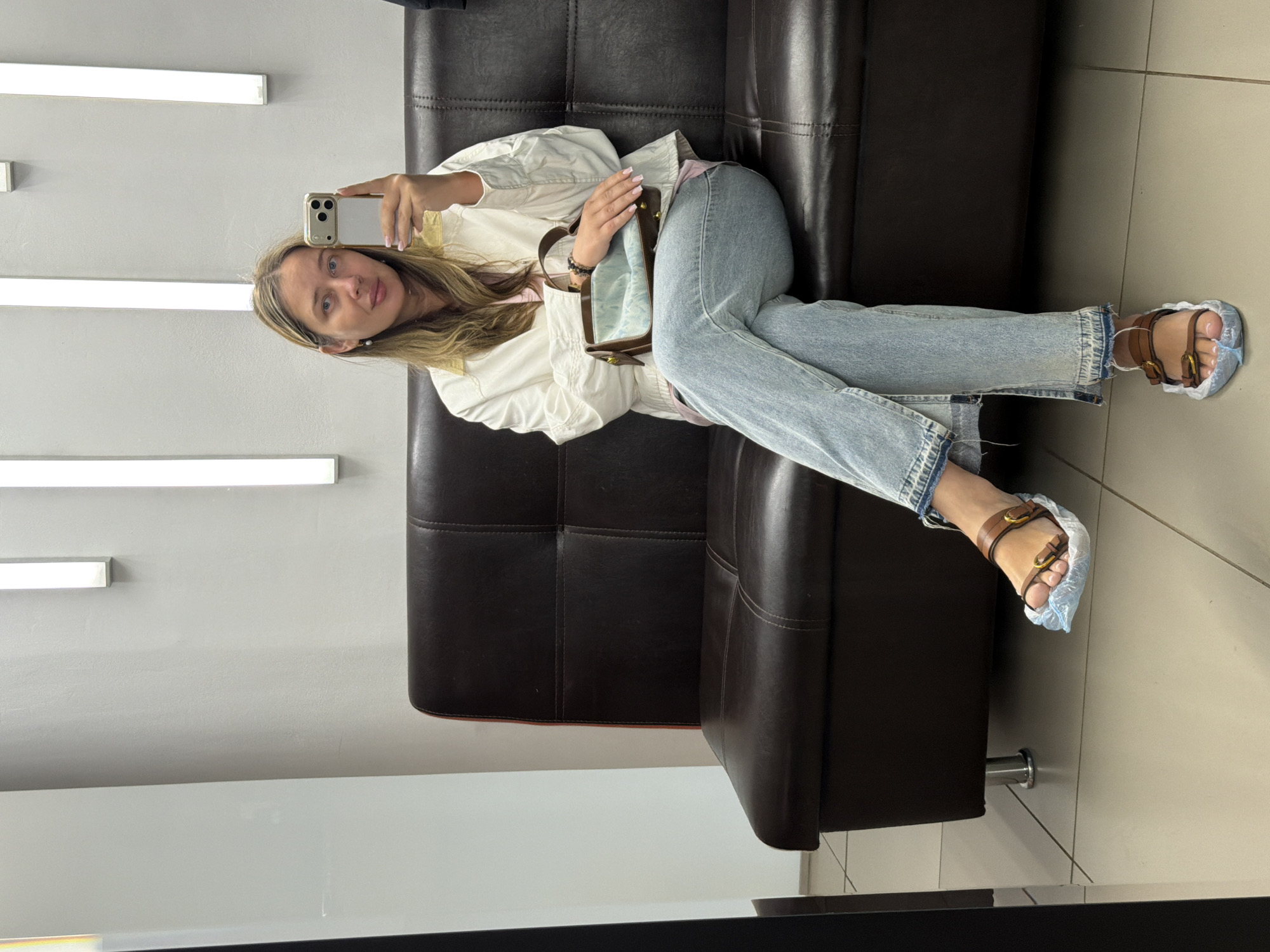 An image captured in an indoor setting, potentially a waiting area in the city of Chaikovsky, Russia, features a single adult female seated on a dark brown, tufted, faux leather couch. She has light-colored hair, visible past her shoulders, and is wearing a white long-sleeved jacket over a light pink top, paired with light blue jeans that have frayed hems and a side slit on the lower leg. Her feet, clad in brown leather sandals with gold buckles, are covered with light blue disposable plastic shoe covers. She holds a light-colored smartphone with a prominent camera array in her right hand, angled as if taking a mirror selfie, partially obscuring her face. Her left hand rests on a small, rectangular handbag with brown leather trim and a light blue interior; a dark beaded bracelet is visible on her left wrist. The couch has silver cylindrical legs. The background consists of light grey walls adorned with three vertical, rectangular LED wall sconces emitting white light. The floor is covered with light-colored square tiles. A dark, vertical reflective surface, possibly a mirror frame or column, is visible on the far left.