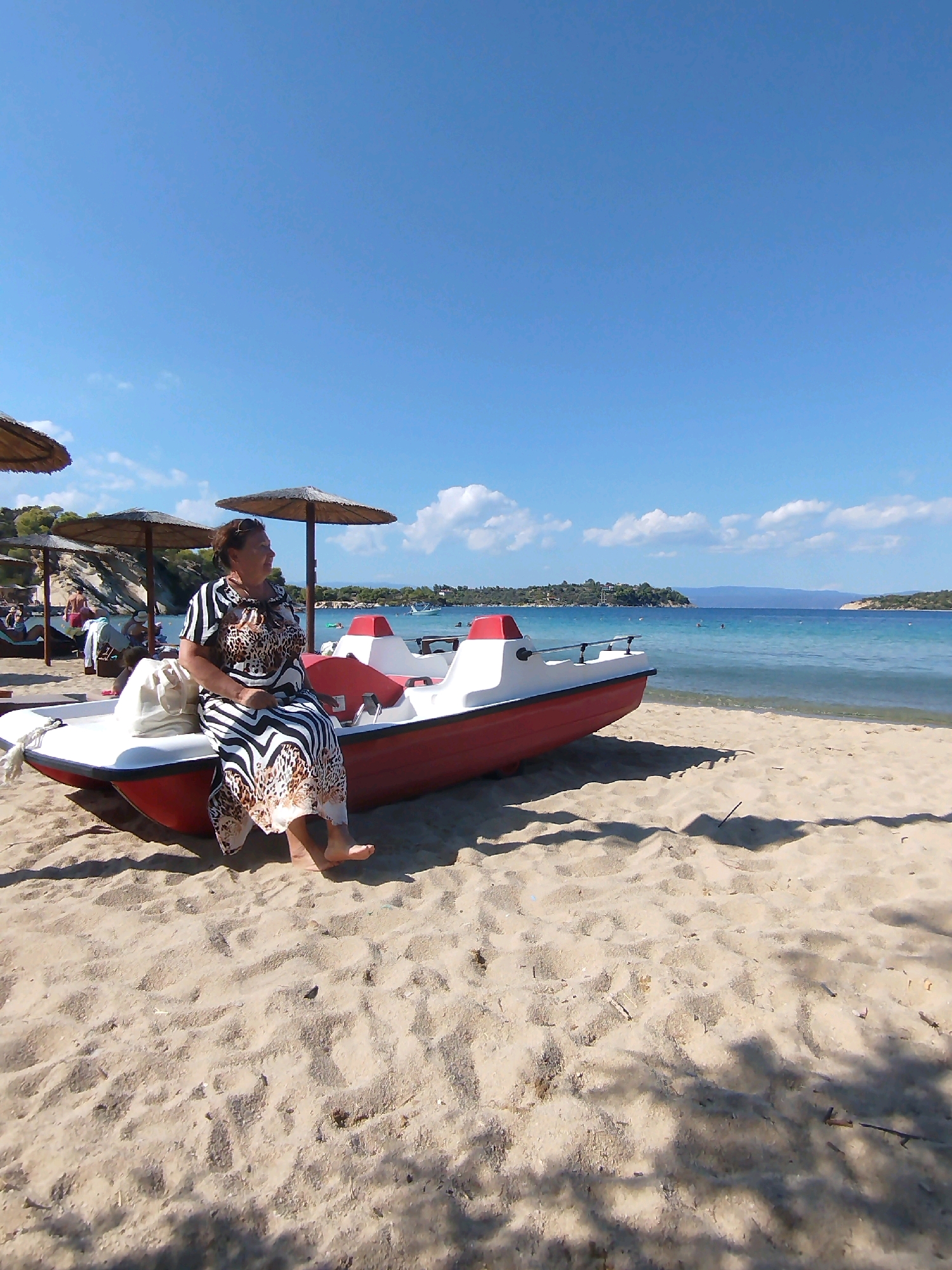 A medium shot captures an outdoor scene on a sandy beach, likely in Sithonia Municipal Unit, Greece, under a clear blue sky. A woman, seated on the edge of a red and white pedal boat, faces away from the camera, looking towards the sea. She is wearing a patterned dress with a zig-zag and leopard print design. A white bag is placed next to her on the boat. The pedal boat is partially on the sand, with its bow facing the left of the frame. In the background, the blue sea stretches out, with a distant coastline featuring green hills and buildings. Several straw umbrellas are set up on the beach. Other beachgoers and lounge chairs are visible in the mid-ground. The sand in the foreground is textured with shadows cast by unseen foliage.