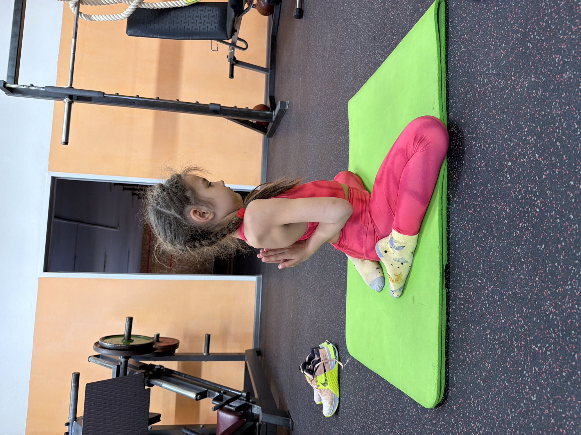 A young girl is performing a yoga pose in what appears to be a gymnasium or fitness center. She is wearing a pink exercise outfit and socks, sitting cross-legged on a bright green yoga mat. Her hands are pressed together in front of her chest. To the left of the girl is a piece of gym equipment holding stacked weights and near her are a pair of athletic shoes. The gym has a gray floor speckled with pink and white and light orange walls.
The geolocation of the image is in the city of Chaikovsky, Russia.