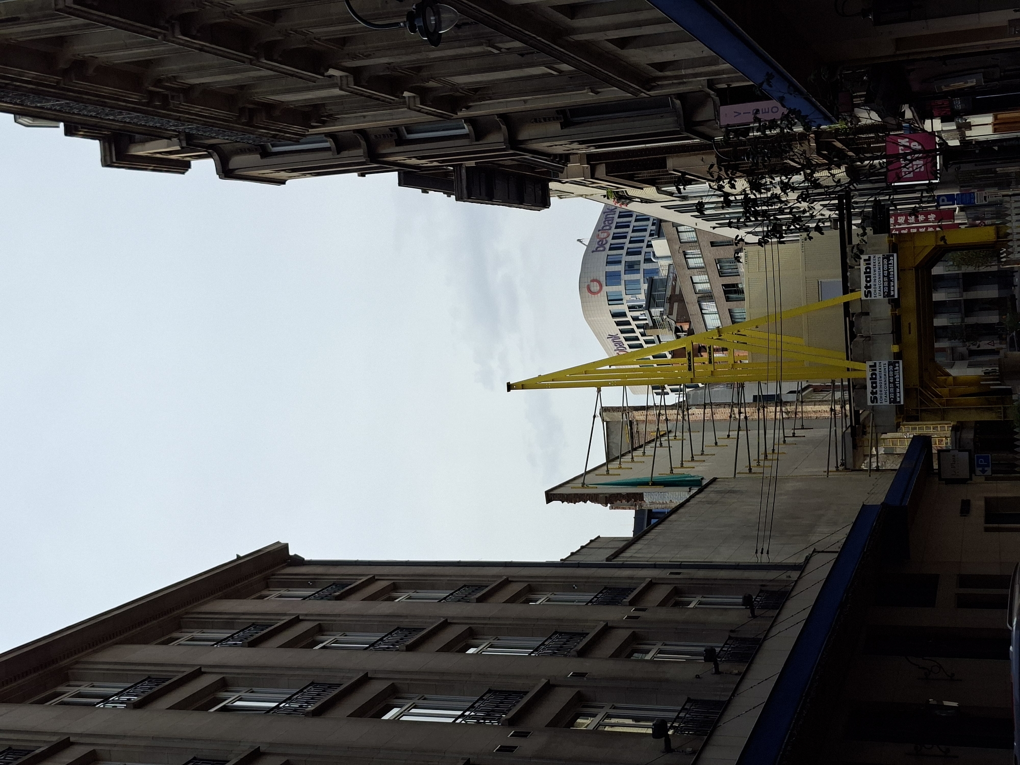 The image depicts a narrow street in Brussels, Belgium, with tall, old stone buildings flanking both sides. In the center of the frame, a bright yellow construction crane is prominently visible, extending upwards and across the street. The crane appears to be part of a larger construction or renovation project, with scaffolding and construction materials evident in the background. Above the crane, a modern, curved white building with the logo "beobank" is visible, suggesting a mix of old and new architecture in the area. The sky is overcast, casting a muted light on the scene. The street itself is lined with shopfronts and entrances, with a few signs visible, including one for "Le Plaza" and another with a "P" symbol, indicating parking. The overall impression is one of a busy urban environment undergoing development.
