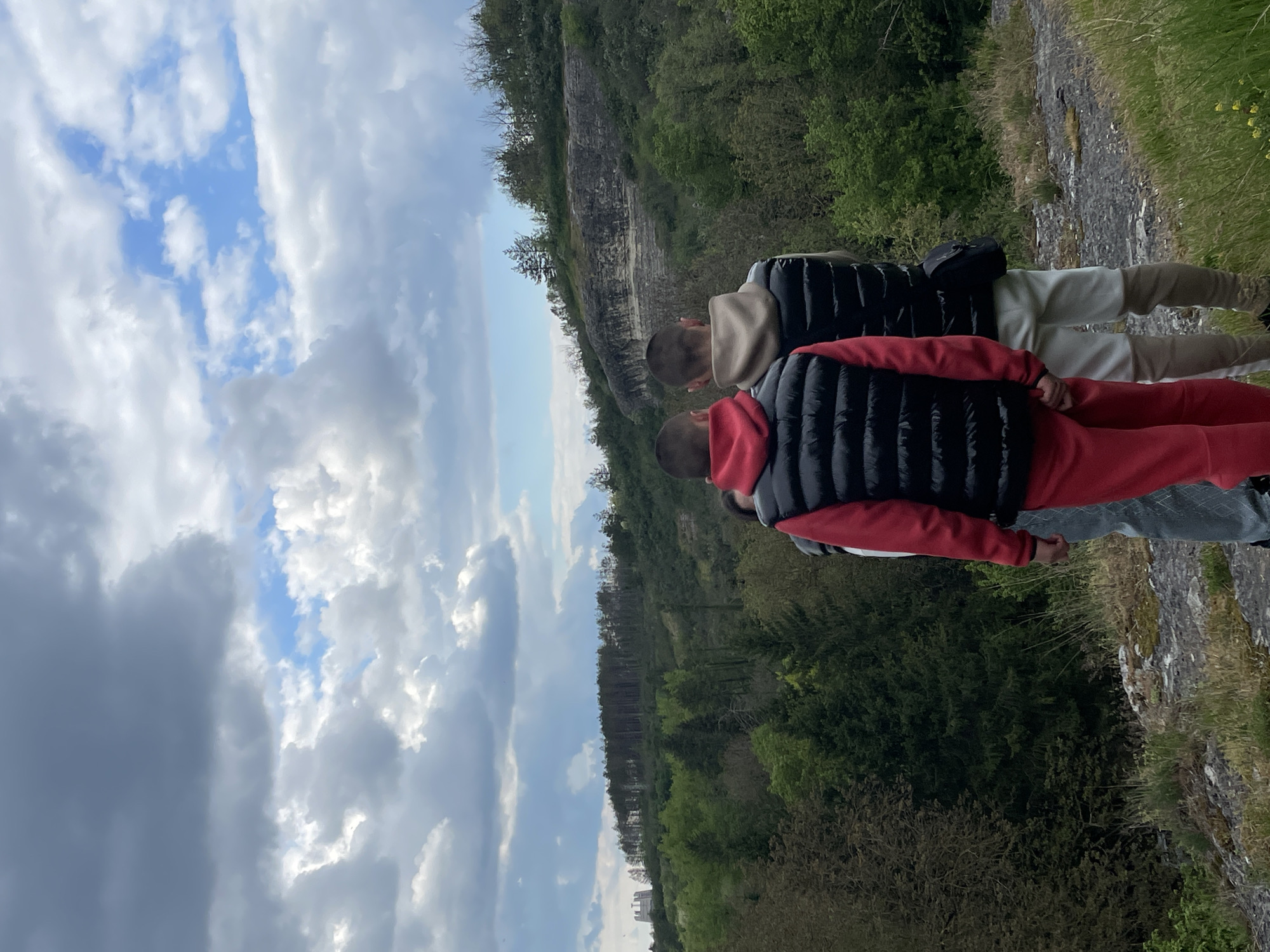 Two individuals are positioned on a rocky edge overlooking a wooded area. The sky above features a mix of clouds and patches of blue. The individuals are facing away from the viewer. The first individual is wearing a red tracksuit and a black gilet. The second individual is wearing a beige tracksuit and a black gilet. The location is Unknown, Unknown.