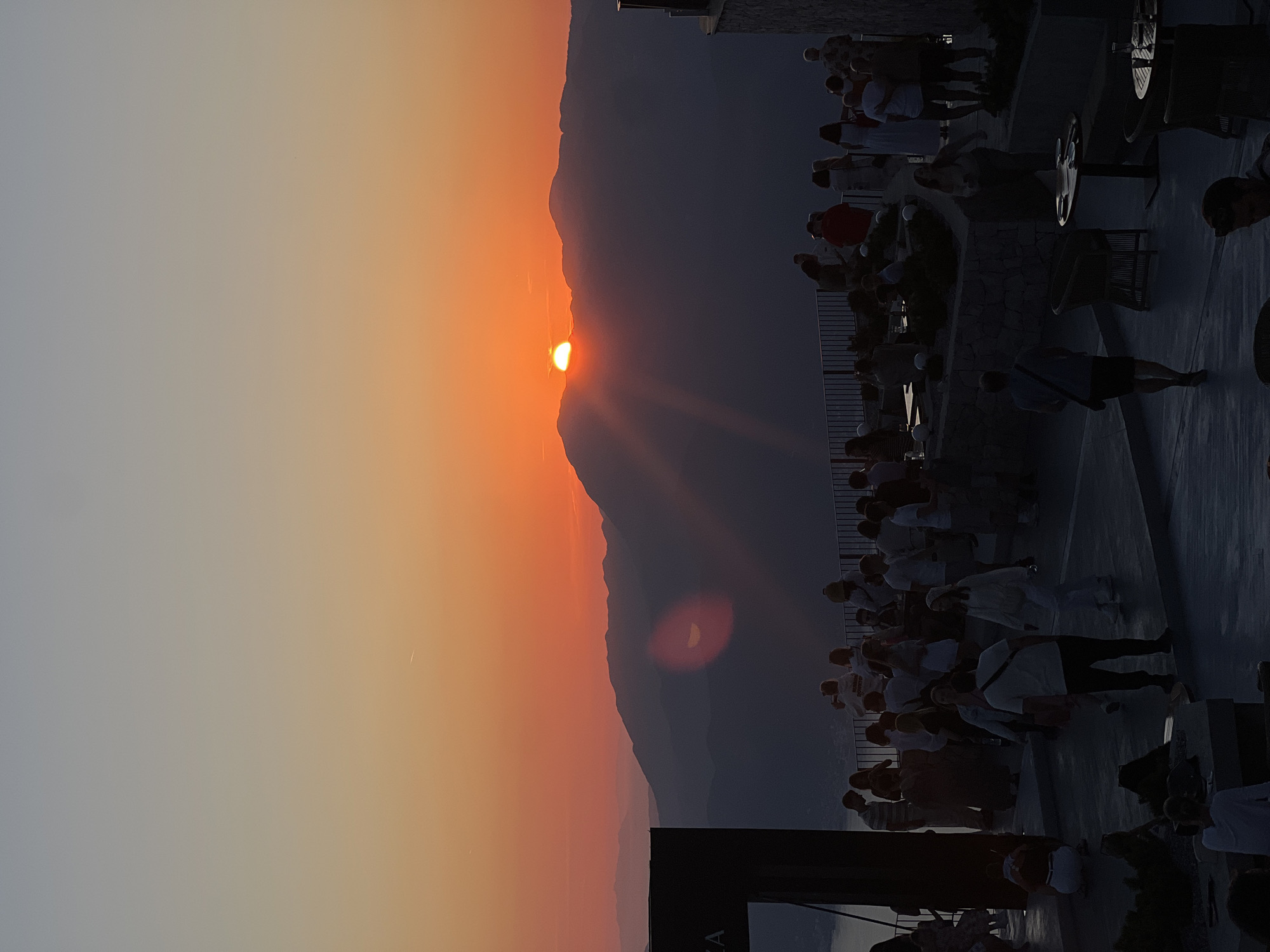 The image displays an elevated outdoor venue, likely a restaurant or bar, at sunset in Kuk, Montenegro. The sky exhibits a gradient, transitioning from light grey-blue at the zenith to vibrant orange and red tones near the horizon. A bright, circular sun disk is partially obscured by the silhouette of a prominent mountain range across the midground, emitting visible light rays. Multiple layers of mountains are visible, presenting as dark silhouettes against the illuminated sky, with distant peaks appearing lighter due to atmospheric haze. A narrow segment of water is visible in the lower-left background. A faint, white linear contrail is present in the upper sky.
The foreground consists of a paved, multi-level terrace, featuring dark metal railings at its edges and retaining walls constructed from cut stone. A large, dark, rectangular architectural structure, possibly a signage panel, is positioned on the far left, with the letters "ZA" partially visible. Spherical white decorative lights are mounted intermittently on the stone walls.
Numerous individuals, estimated to be several dozen, populate the terrace. Their actions include standing, walking, conversing in groups, and observing the sunset. Several people are holding mobile devices, ostensibly for photography. One individual in the mid-ground is wearing a white garment with the text "RUNNING BOUNDARY." In the lower foreground, a person walks away from the viewer, carrying a dark cross-body bag. Other individuals are seated at round tables furnished with wicker and standard outdoor chairs. Drinking glasses are discernible on some tables. A distinct red, crescent-shaped lens flare is visible in the mid-left portion of the image. The attire of the people appears casual to semi-casual, consistent with an outdoor evening setting.