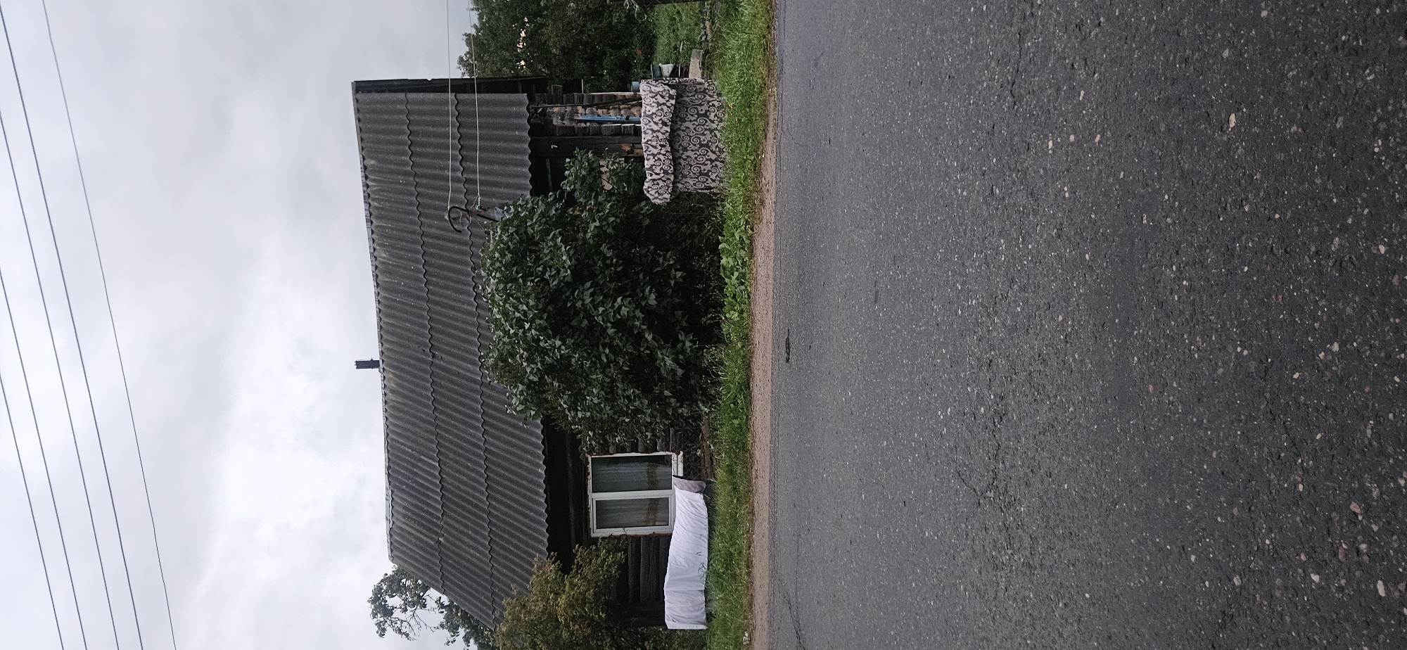 In the city of Liozna, Belarus, a weathered wooden house stands beneath a cloudy sky. The house has a dark, corrugated roof and a single window with white trim. To the left of the house, a white sheet is draped over a low structure, possibly for drying clothes. A large, dark green bush obscures the right side of the house. In front of the house, a well is covered with a patterned fabric, featuring swirling designs. The foreground is dominated by a grey asphalt road with some cracks and scattered pebbles. Power lines are visible in the sky above the house. The scene appears to be a rural setting.