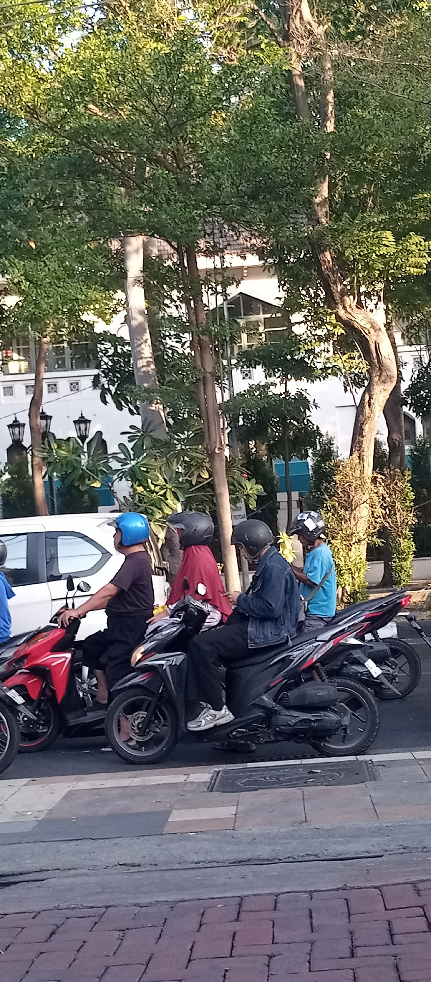 The image depicts a street scene in Surabaya, Indonesia, with several individuals riding motorbikes. In the foreground, a man wearing a blue helmet and a dark t-shirt is on a red motorbike. To his right, a person in a red headscarf and dark clothing is on a black motorbike, followed by a person in a denim jacket on another black motorbike. Another person in a light blue shirt is visible on a motorbike further to the right. The background shows a white building with traditional architecture, surrounded by lush green trees and street lamps. The ground is paved with bricks and concrete. The lighting suggests it is daytime, possibly late afternoon, with sunlight casting shadows.