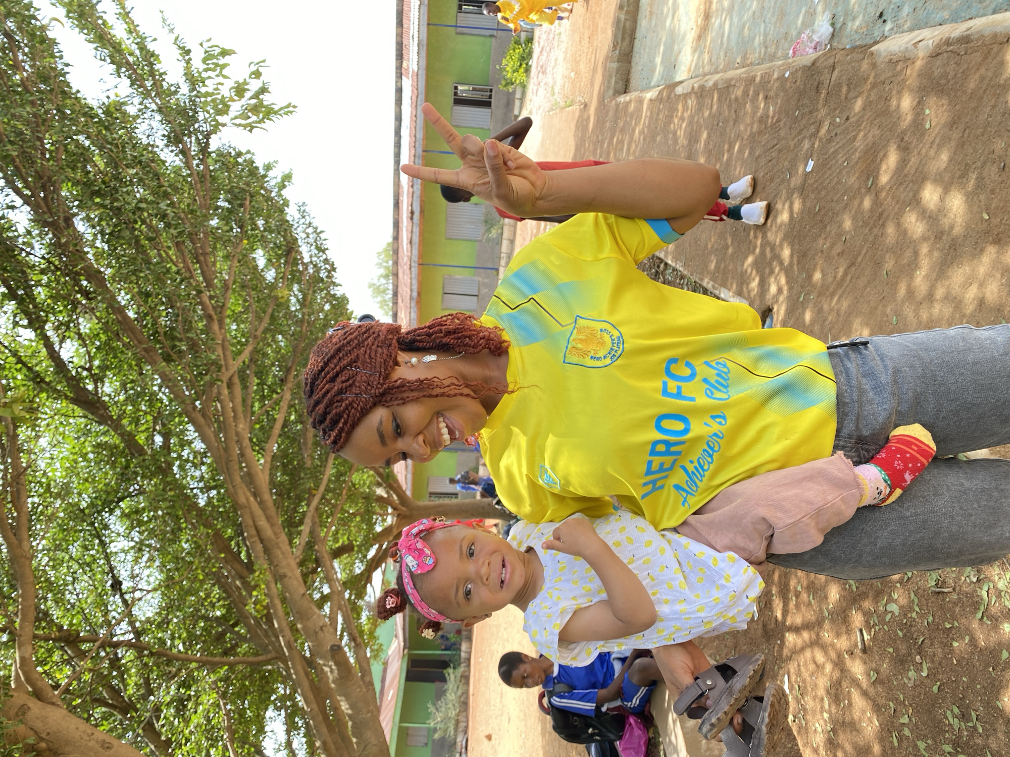 A woman in a yellow t-shirt and jeans is holding a young child in her lap outside in Gura Top, Nigeria. The woman is smiling and giving a peace sign. The child is wearing a polka dot dress and a pink headband. A tree is on the left side of the image and a building is in the background. Other people and objects are visible in the background.
