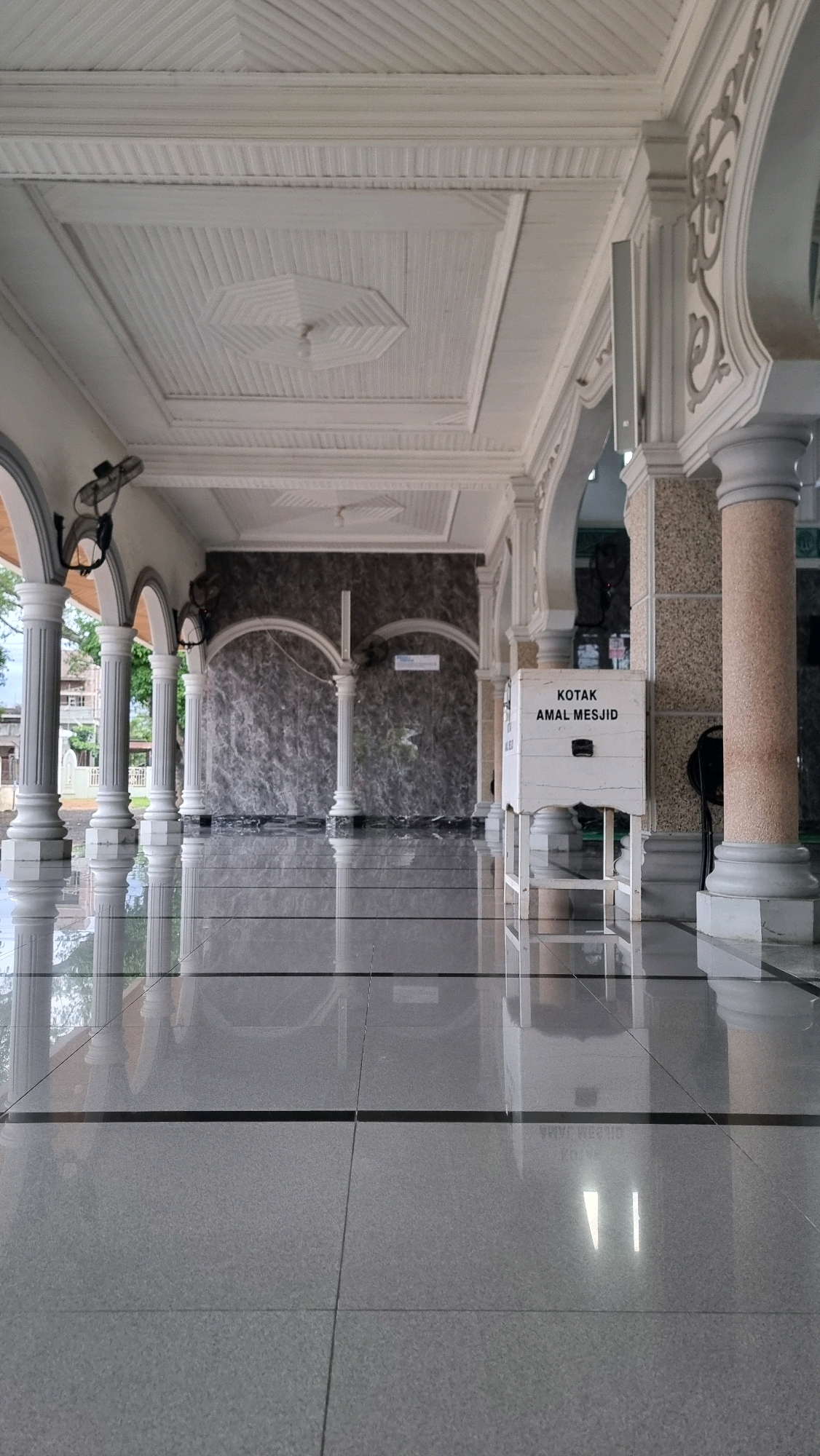 The image shows an indoor hallway with a high, ornate ceiling and polished floors. White pillars line the hallway, supporting arches that lead to a dark, textured wall in the background. On the right side of the hallway, a white donation box with text in a foreign language is visible, placed on a stand. Two ceiling fans are mounted on the wall. The floor is made of large, grey tiles that reflect the surrounding architecture. The environment appears to be a building in Manetunong, Indonesia.